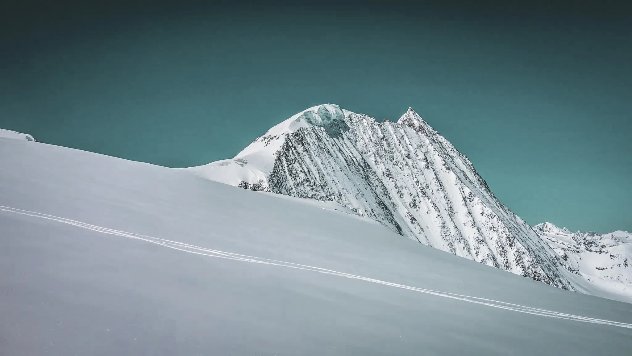 A majestic mountain peak, covered in immaculate snow, rises up against a deep blue sky. The steep slopes are criss-crossed with lines that suggest ski tracks, a testament to the mountain's past.