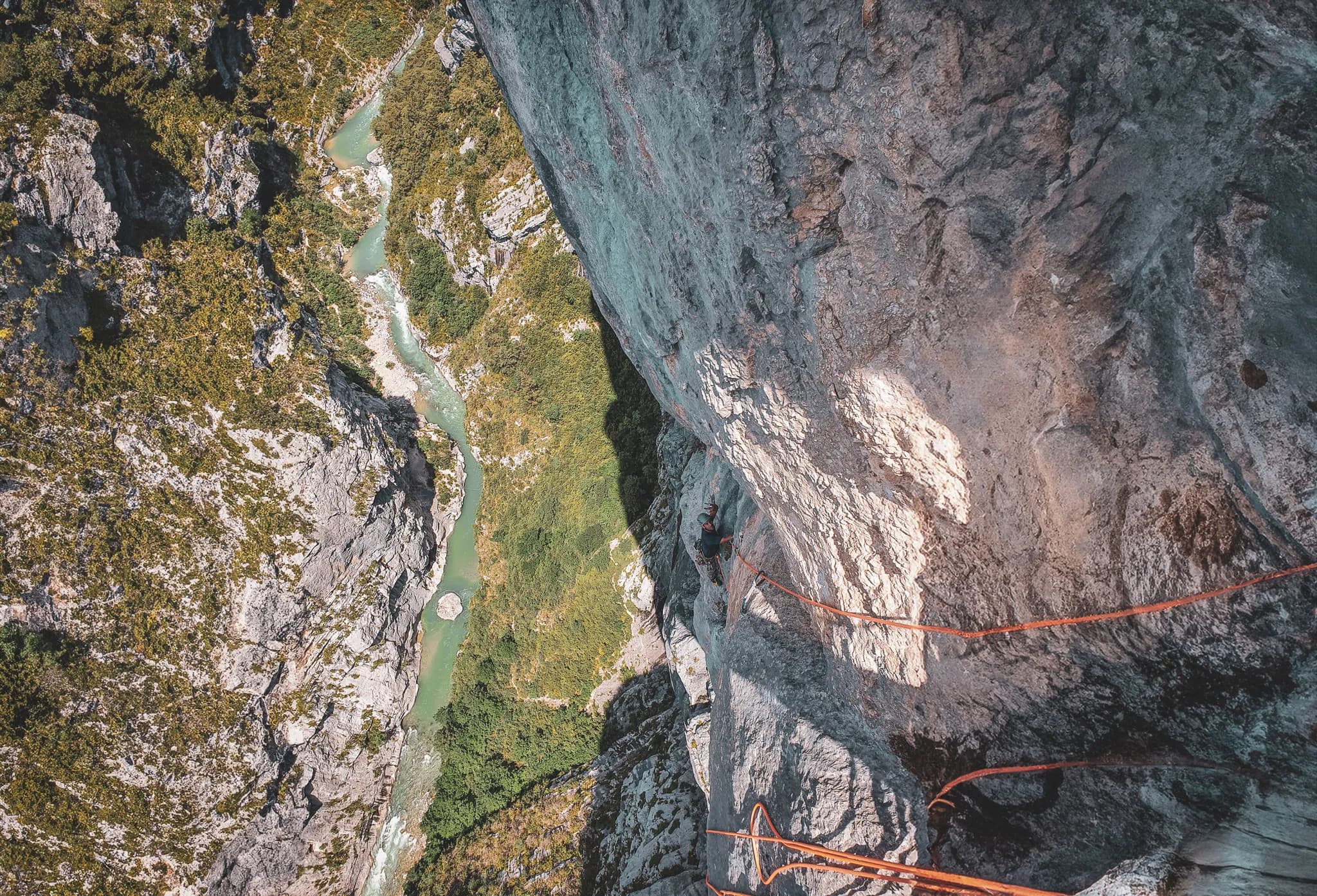 A climber ascends a vertical rock face, surrounded by cliffs and lush vegetation. Below, a meandering river with turquoise waters cuts through a verdant landscape, while the texture and nuances of the