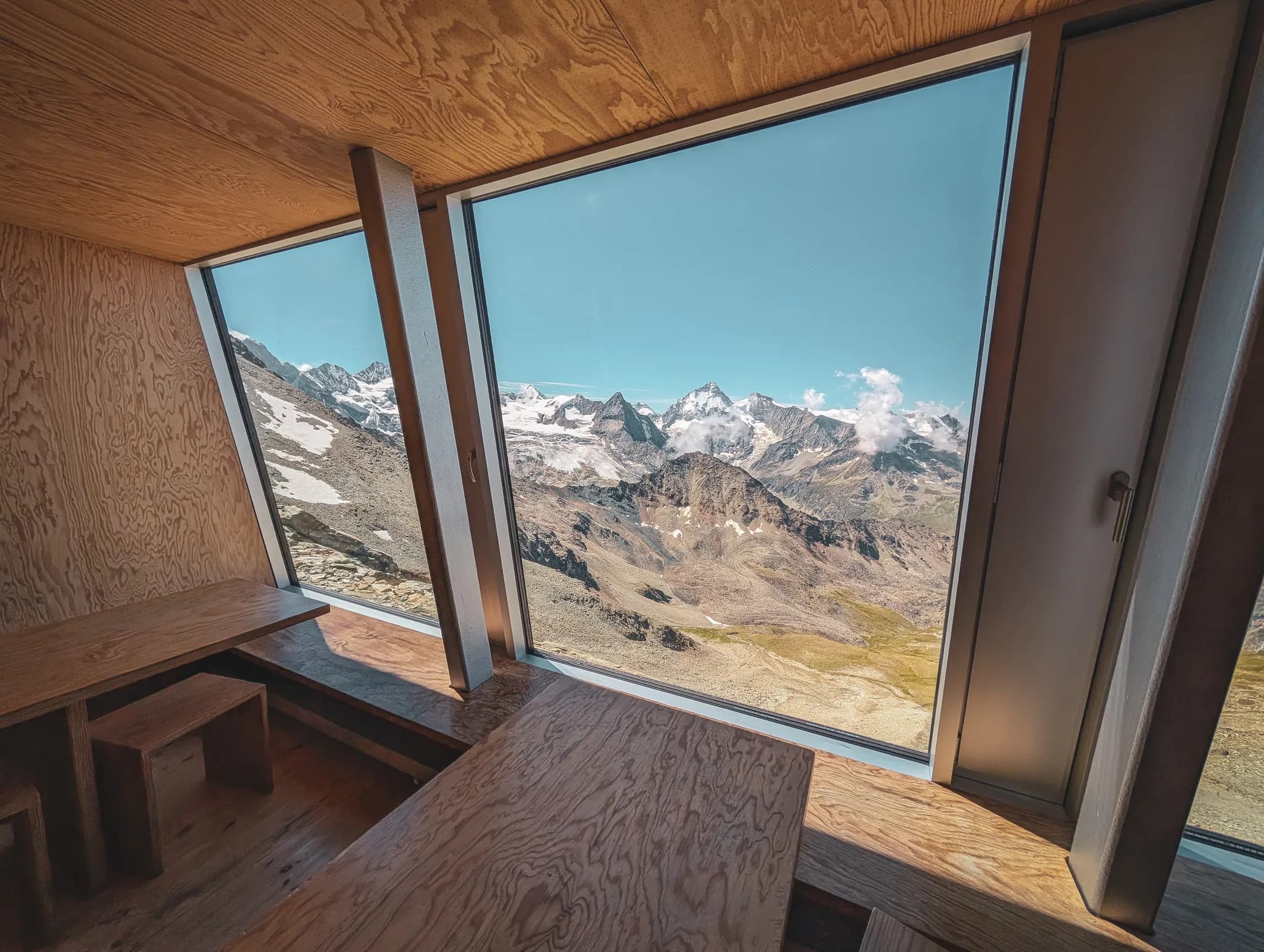 A modern building in the mountains, perhaps a Mountain hut or chalet, is surrounded by majestic Alpine scenery. People are gathered outside, admiring the view, while a Swiss flag flutters in the wind.