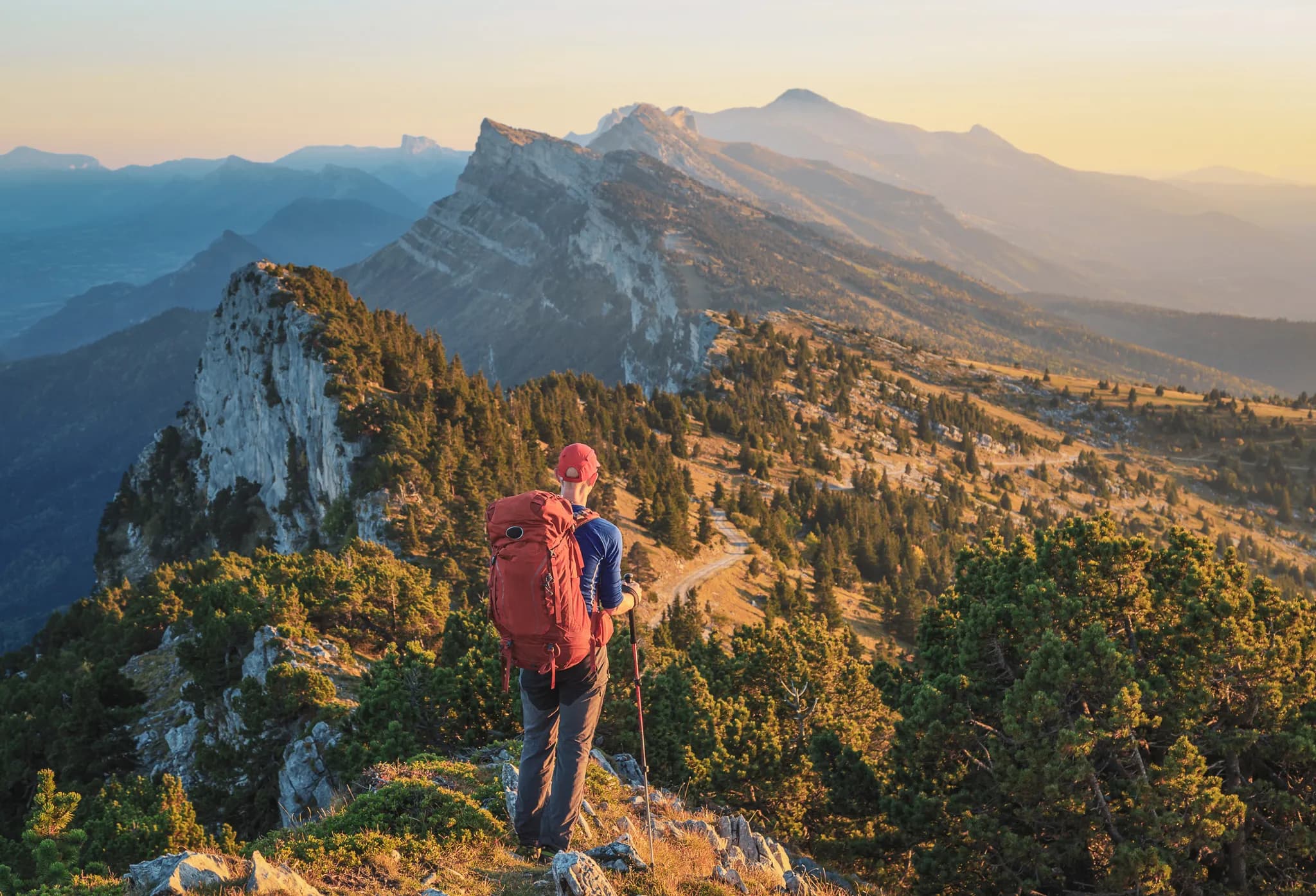 Un randonneur se tient sur un sommet montagneux, admirant un vaste paysage de collines et de montagnes sous un ciel lumineux au crépuscule. L'individu, portant un grand sac à dos rouge et un chapeau, regarde vers