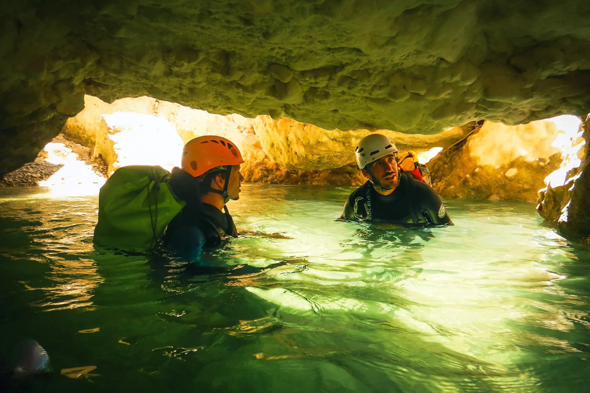 Two people in an aquatic environment, immersed in a cave with clear water. They are wearing safety helmets, one orange and one white, and diving suits. One of the adventurers