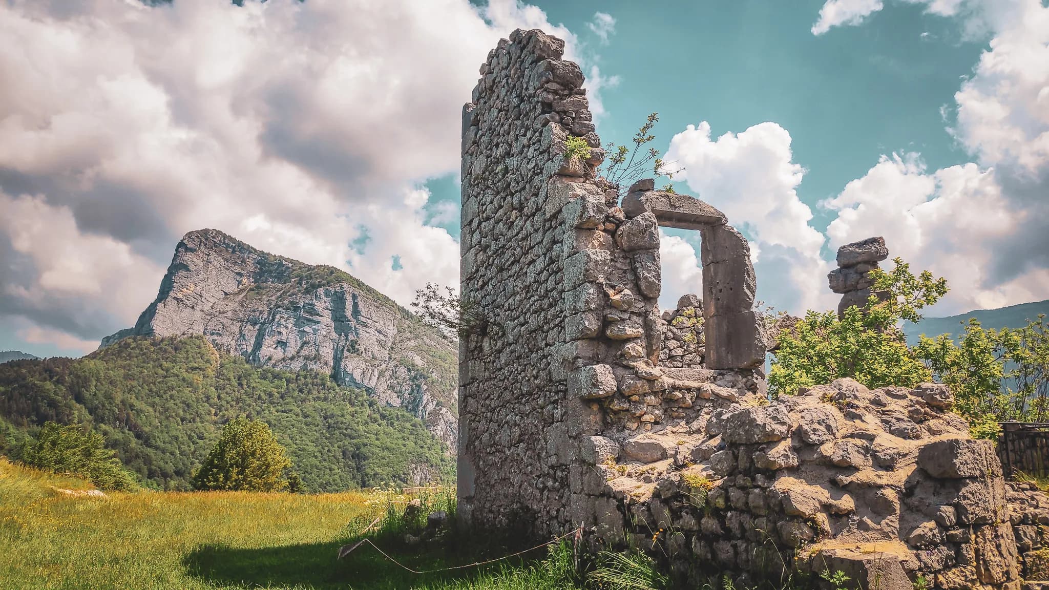 Des ruines en pierre d'un ancien bâtiment se dressent au milieu d'une verdure luxuriante, avec une grande montagne rocheuse en arrière-plan. Le ciel est parsemé de nuages blancs et moelleux. La lumière du