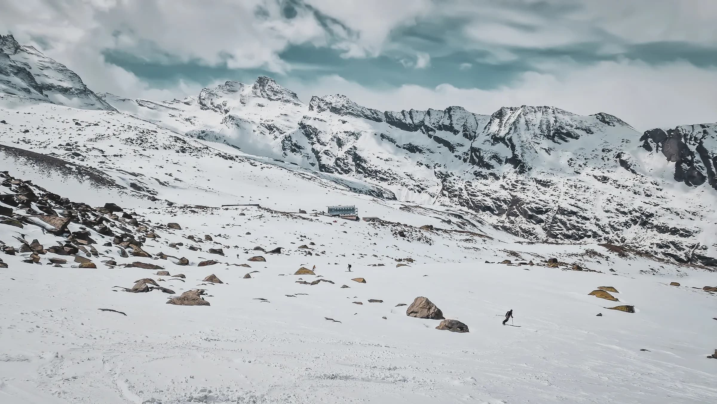 A snowy landscape with majestic mountains in the background, covered in snow and partially obscured clouds. Scattered boulders cover the ground, while a building rises out of the sky.