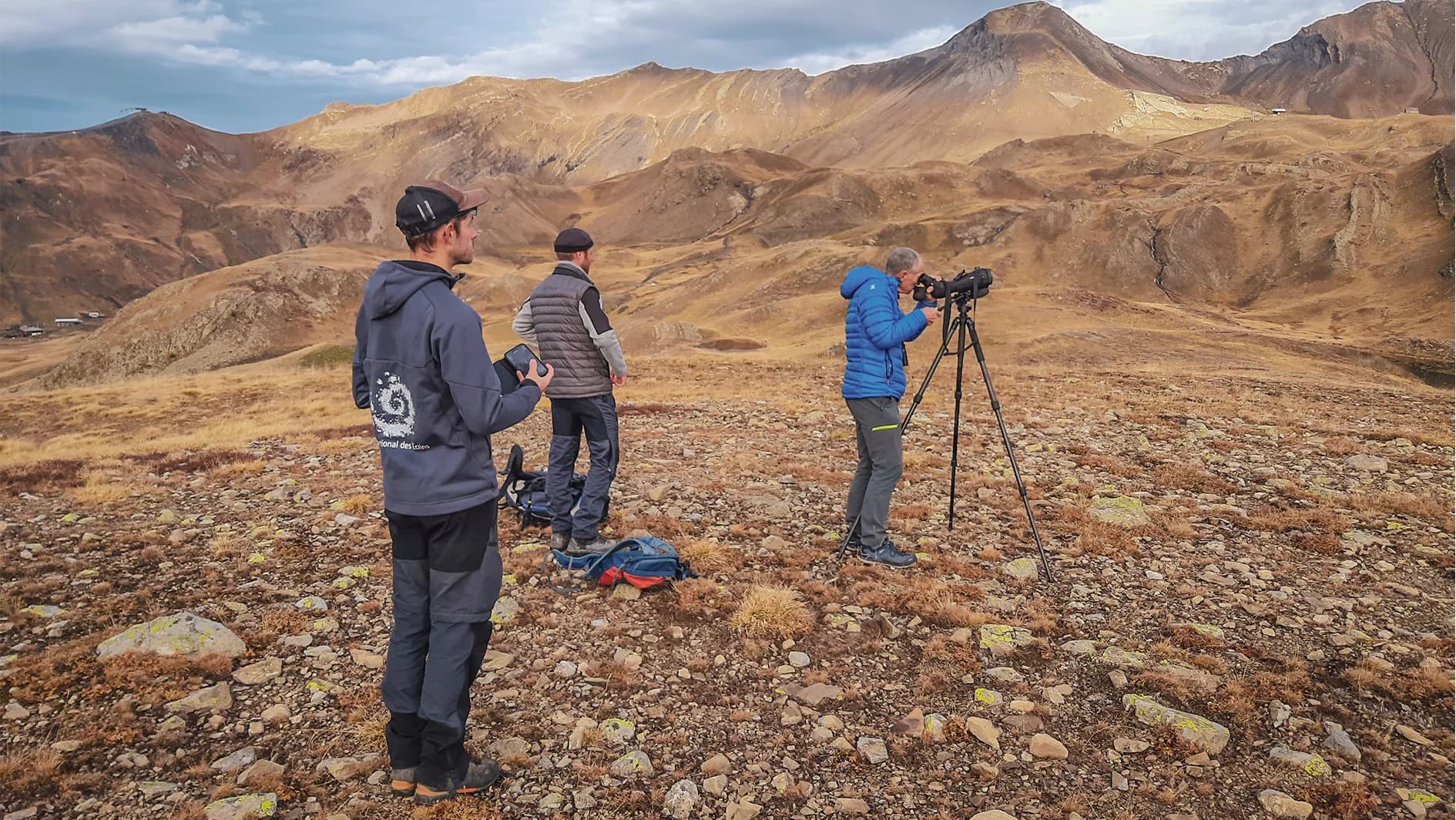 Three people stand on rocky ground, surrounded by mountains of golden and brown hues. A man at the back, wearing a grey jumper, looks out over the landscape, while another, wearing a blue jacket