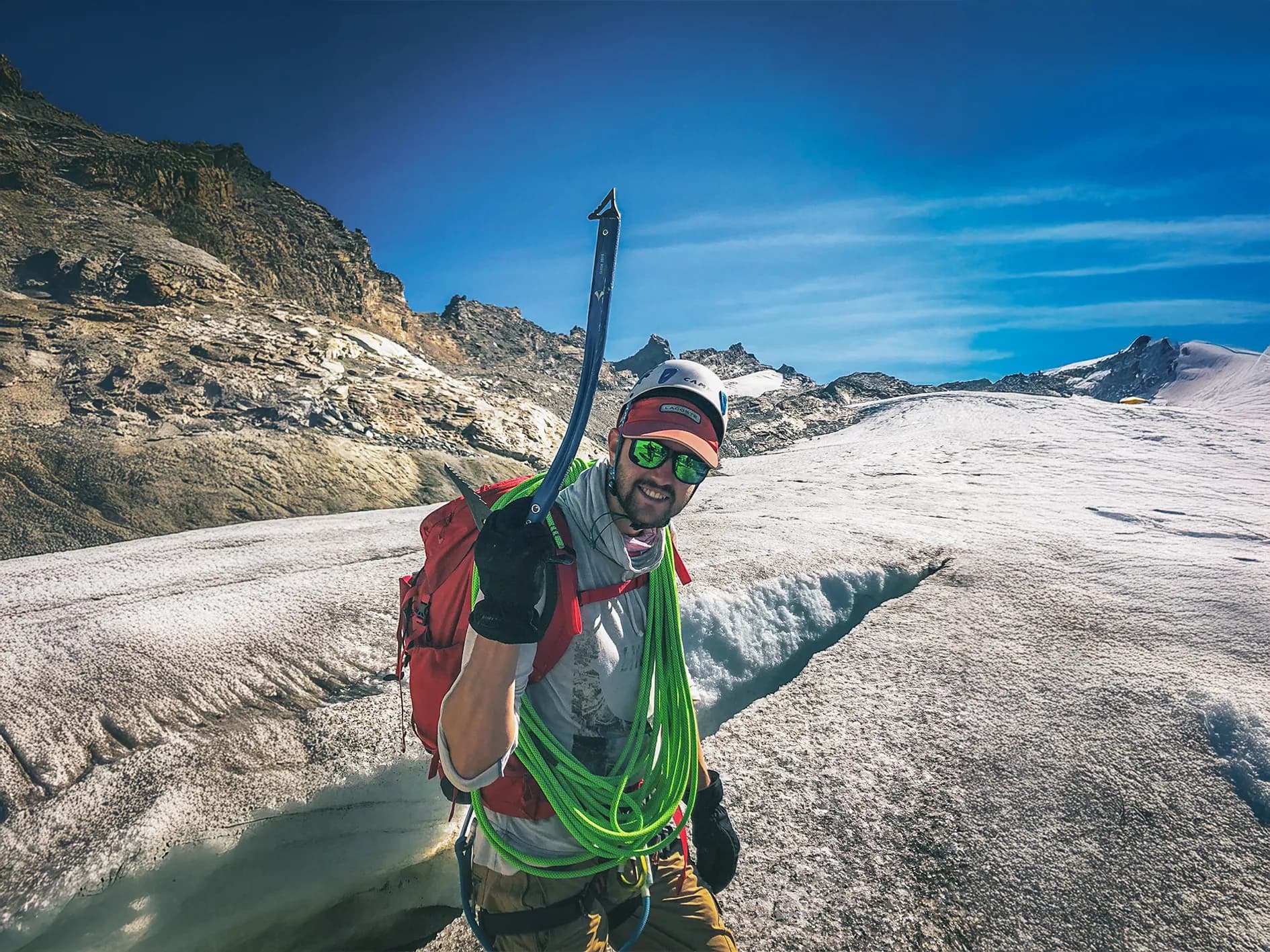 A mountaineer stands on a glacier, smiling and brandishing an ice axe in one hand. He is wearing a red cap, reflective sunglasses and black gloves. On his back, a rucksack