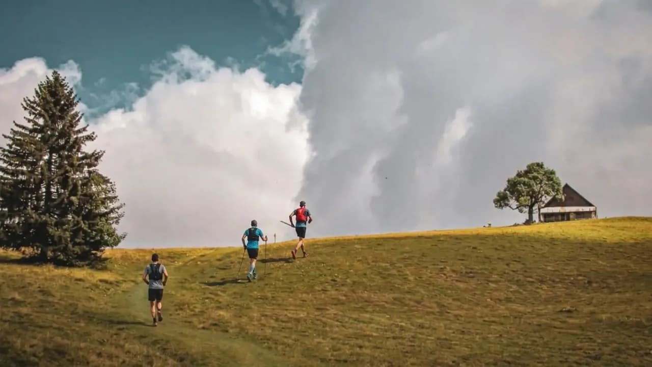 Trois randonneurs s'élancent sur une colline verdoyante, avec des nuages majestueux en arrière-plan.