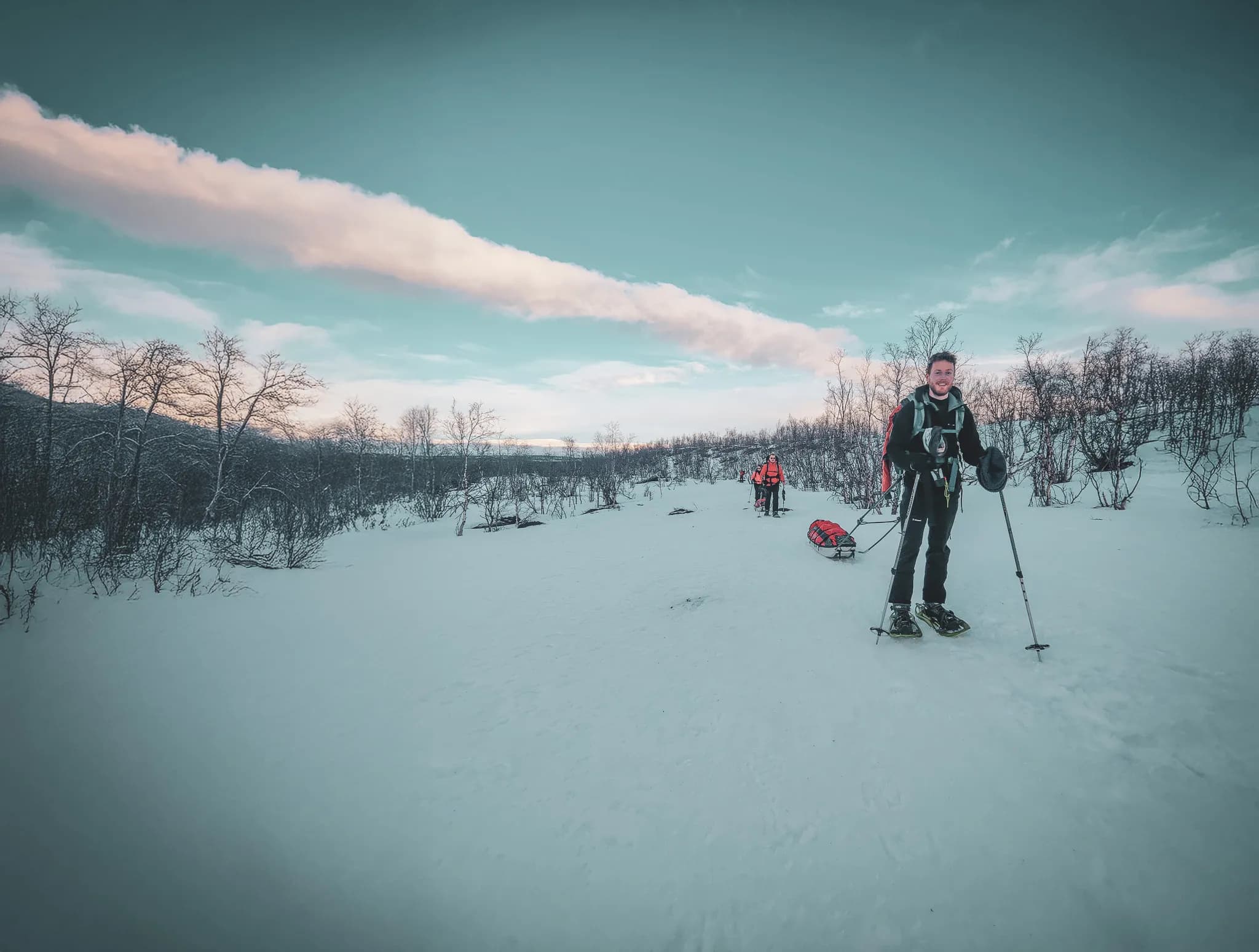 A snowy landscape with bare trees in the background. Two people come forward, equipped for winter walking. One of them, smiling, is wearing snowshoes and holding poles.