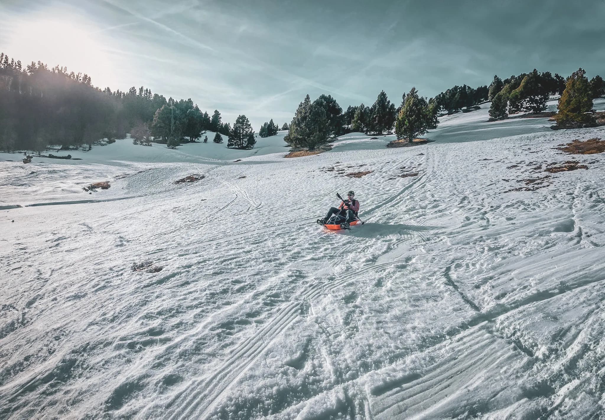 A snowy landscape with a person sitting on an orange sledge, gliding across a wide expanse of snow. The scene is surrounded by fir trees and hills. The sky is clear with a soft light, suggesting