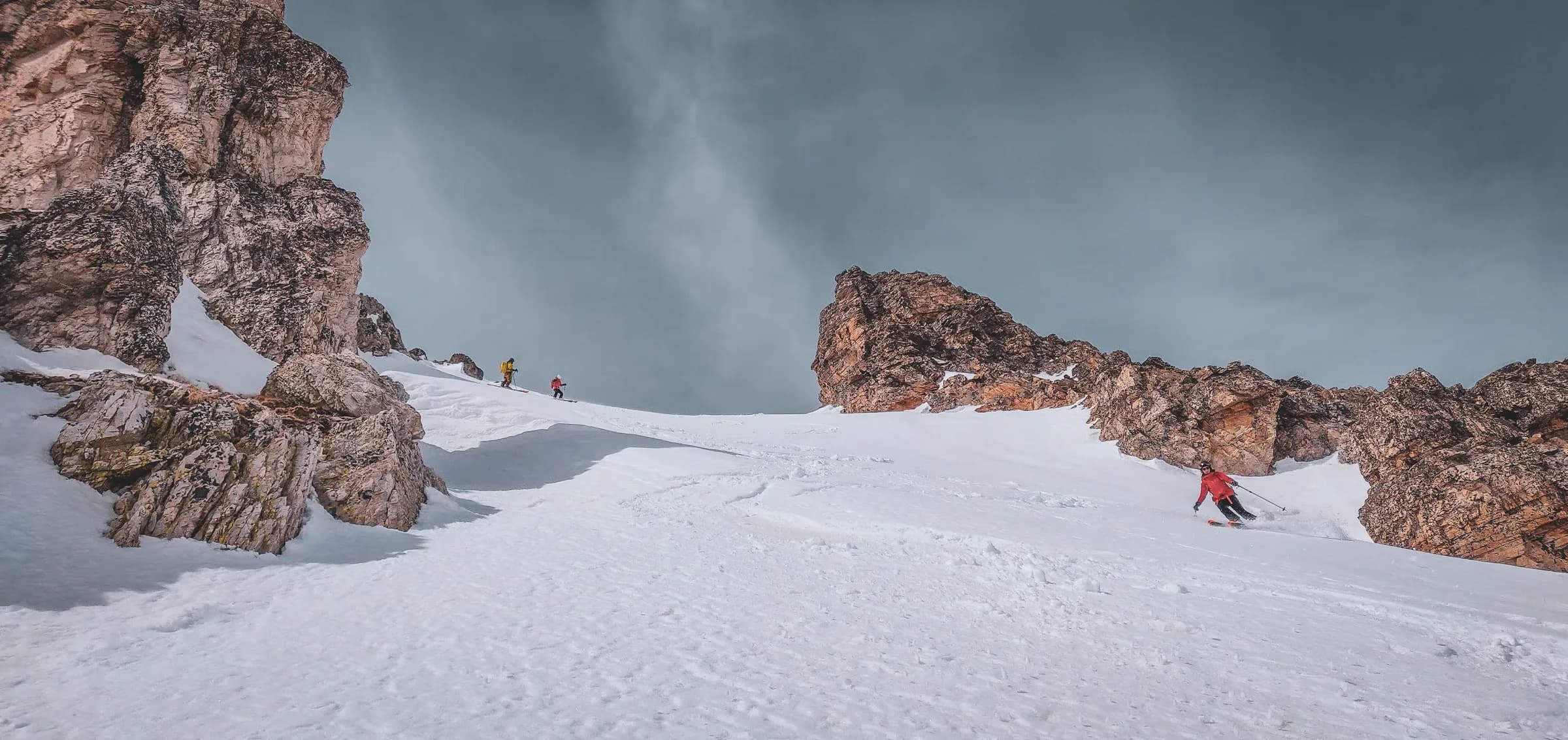 Un paysage hivernal en montagne, caractérisé par des flancs enneigés et des rochers dénudés. Deux skieurs, habillés de vêtements colorés, descendent un versant recouvert de neige, tandis qu’une