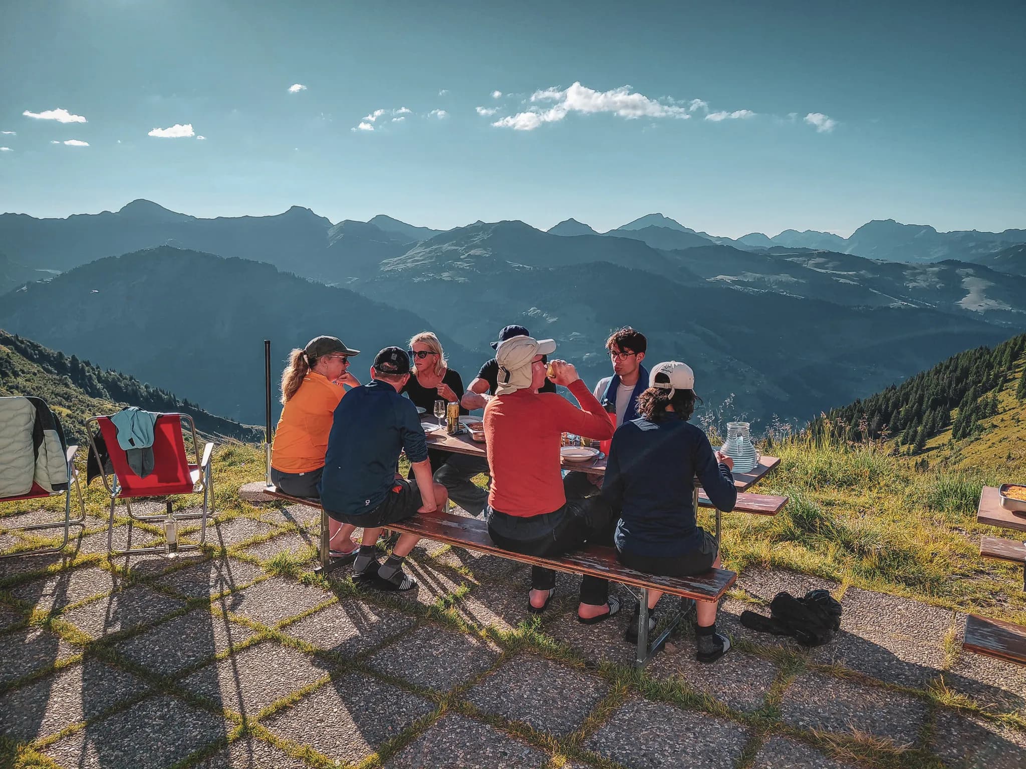 A wooden hut, typical of the mountains, is surrounded by majestic mountains and greenery. Picnic tables adorned with colourful parasols welcome visitors enjoying the panoramic views.