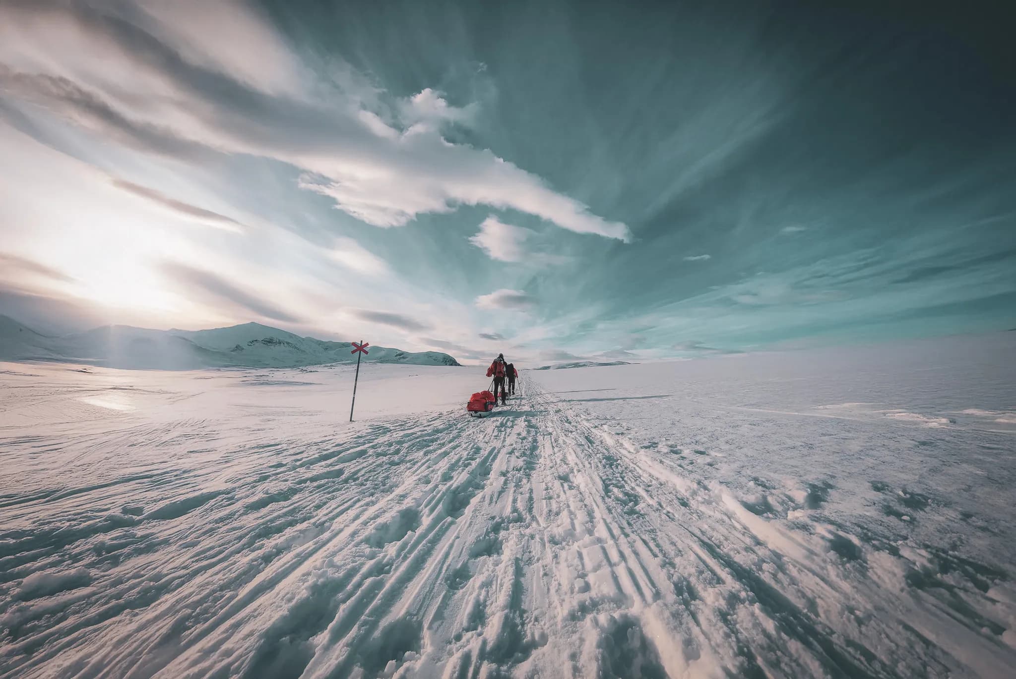 The snow-covered, signposted Kungsleden trail stretches out under a cloudy sky, with wisps of cloud stretching to the horizon. In the centre of the image, two people are pulling sledges.