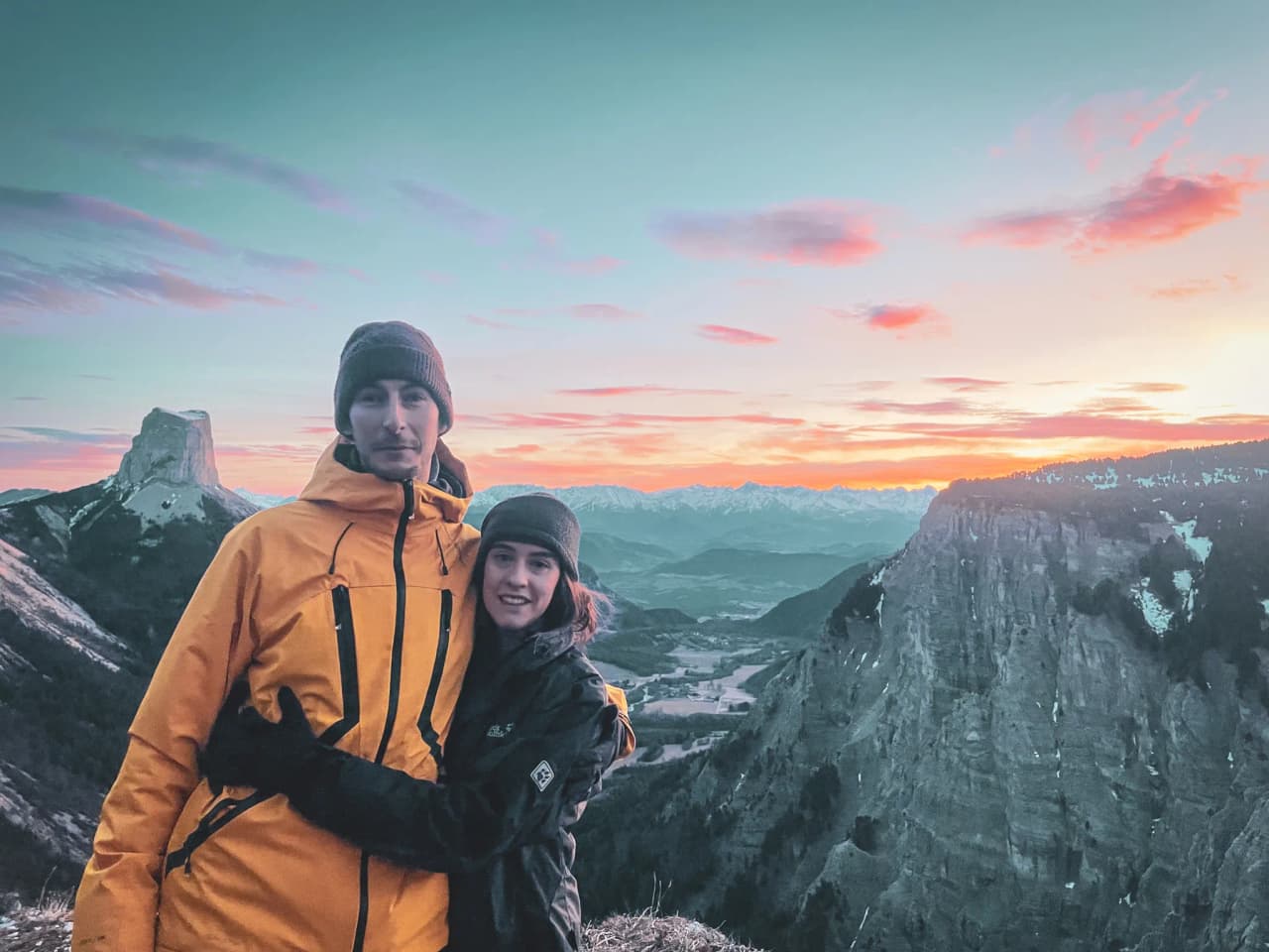 A smiling couple in the mountains, dressed in winter clothes, with a magnificent sunset in the background.