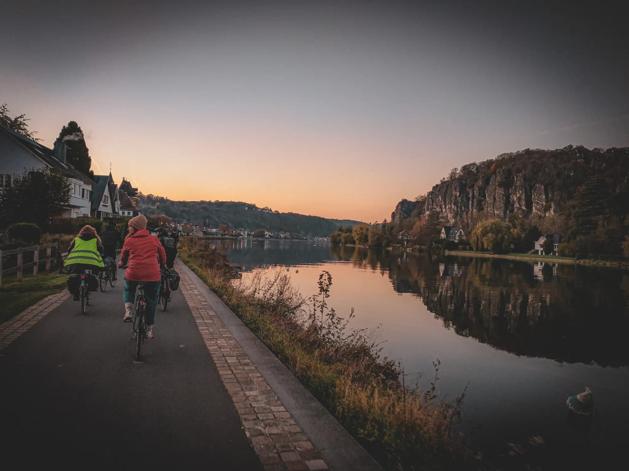 Un groupe de cyclistes se déplace le long d'une piste cyclable bordée de maisons, avec un paysage paisible à leur droite. Leurs vélos sont équipés de sacs, et l'un des cyclistes porte une veste réfléchissante