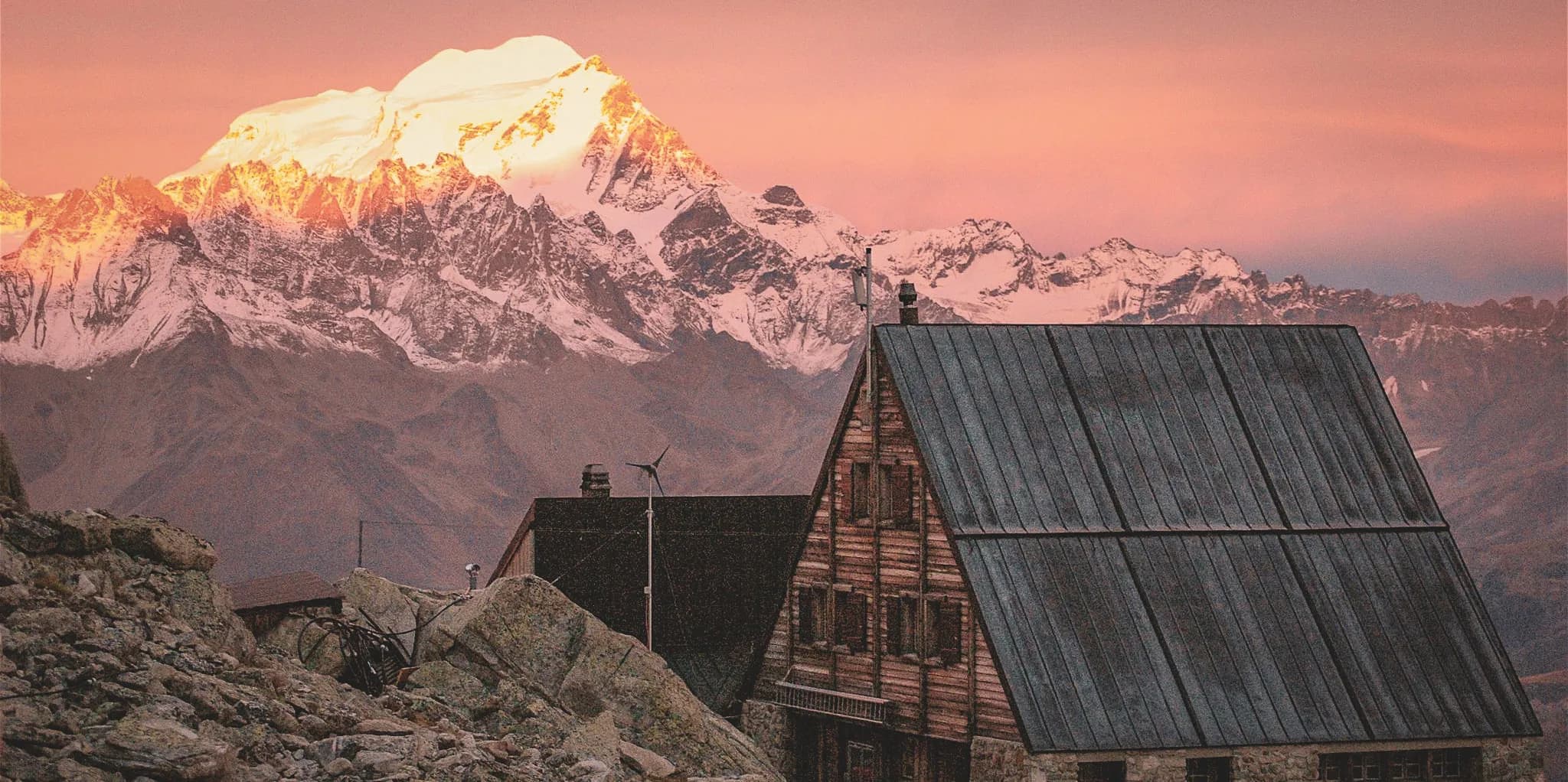 A spectacular mountain landscape at dusk, with a snow-capped peak illuminated by the golden light of the setting sun. In the foreground, a charming wooden building with tin roofs, blending harmoniously into its surroundings.
