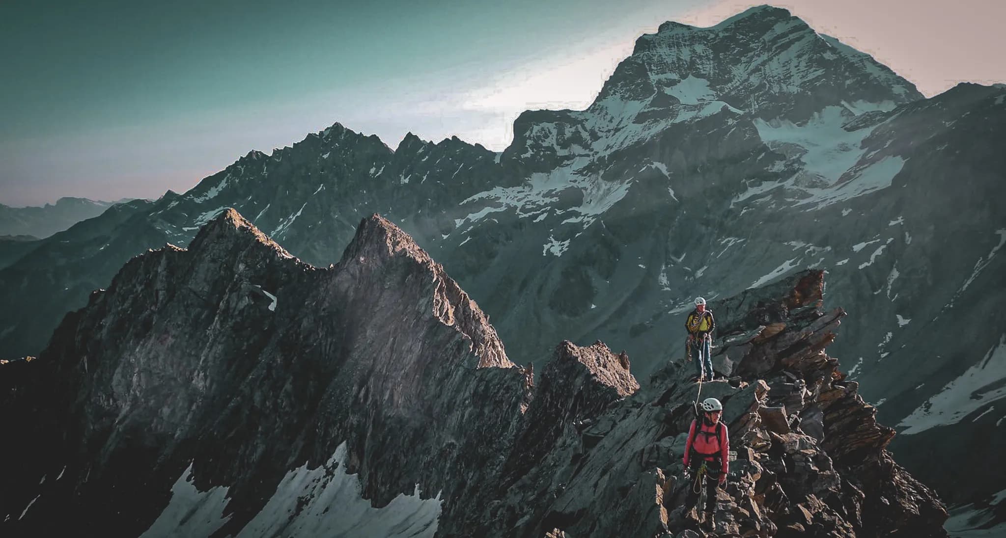 A spectacular mountain landscape with steep, snow-capped peaks. Two climbers, equipped with safety gear, climb a rocky ridge, adding to the sense of challenge and adventure.