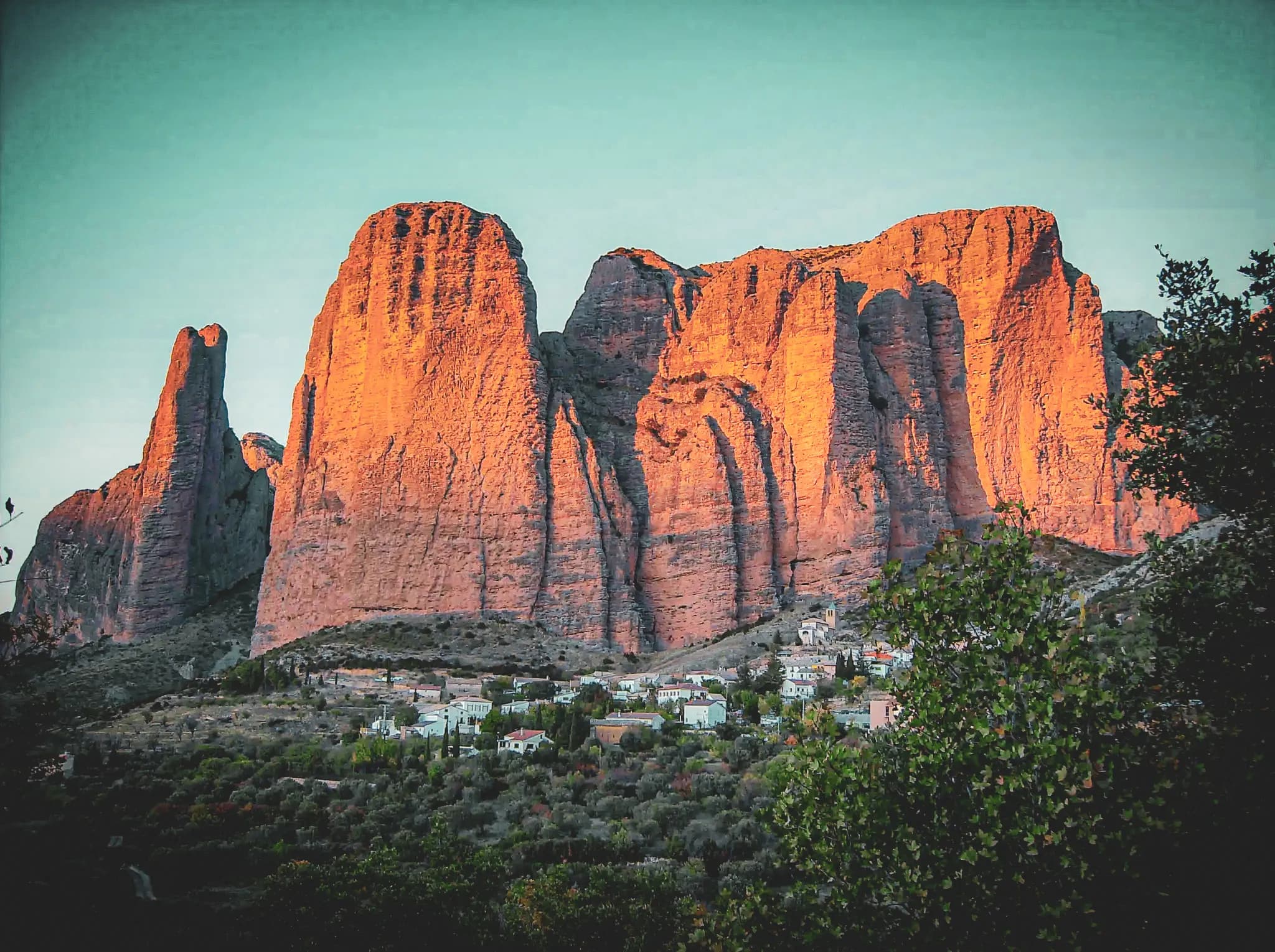 Majestic cliffs in shades of orange dominate the landscape, illuminated by the sunlight. At the foot of the mountains, a small village with white houses lies amid lush greenery.