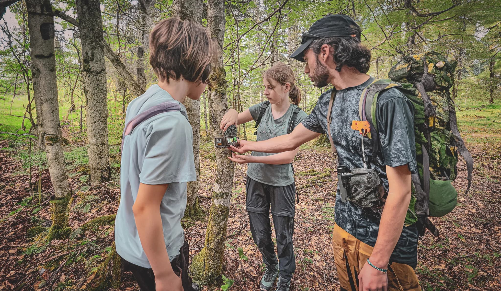 Three people in the great outdoors examine a camera trap on a tree, surrounded by lush greenery.