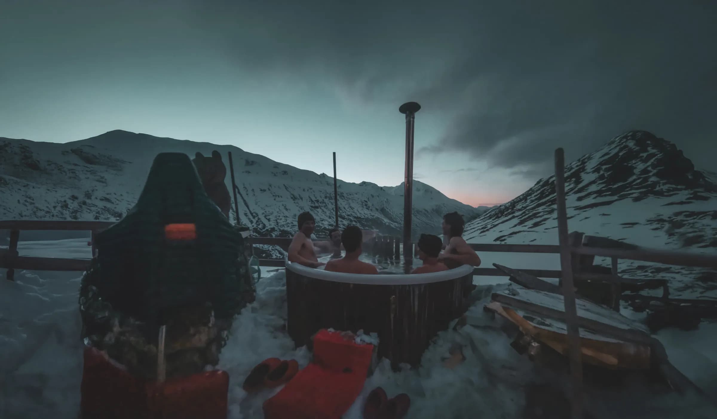 A group of people are in a wooden hot tub outside, surrounded by snow and mountains. The atmosphere is peaceful, with the sky turning from blue to pink at dusk. Visit