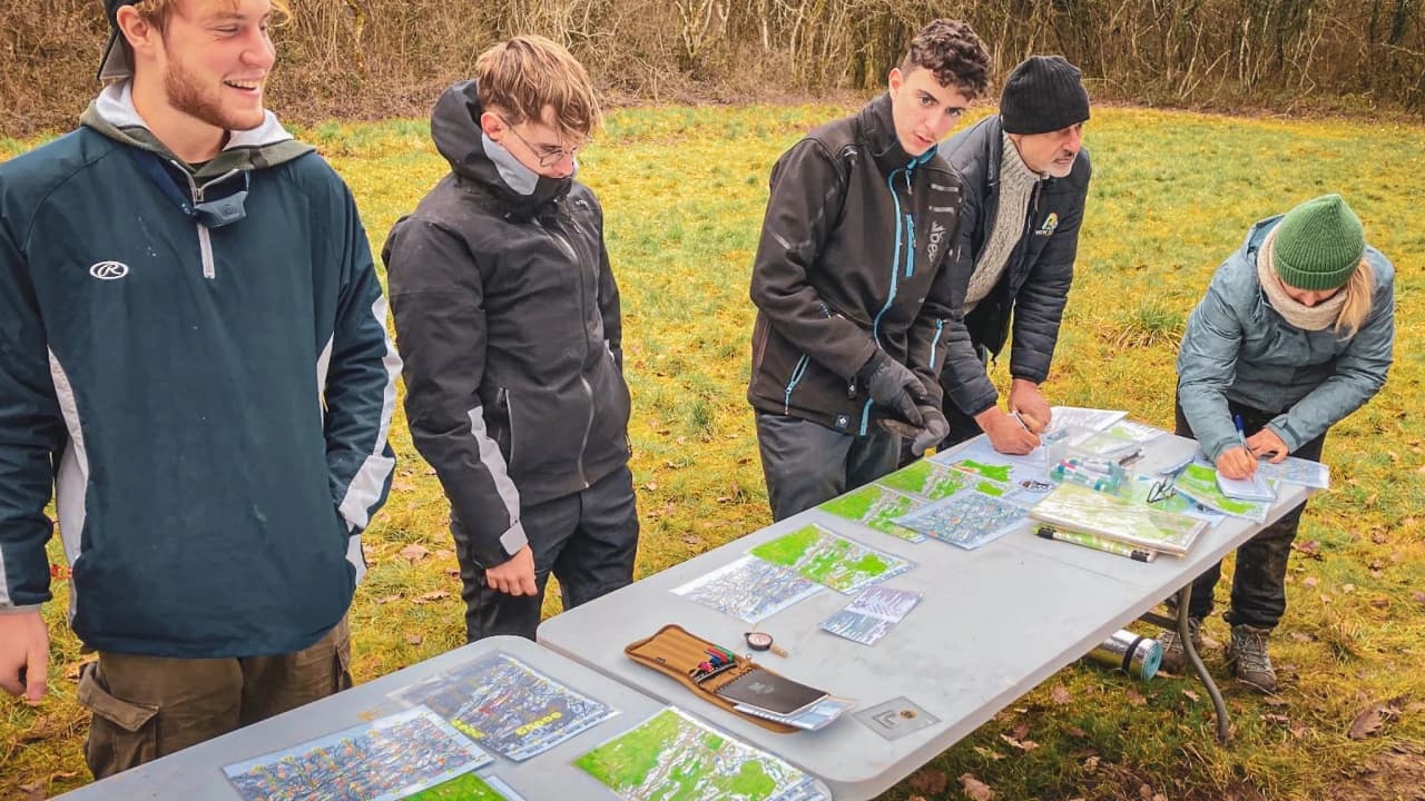Cinq personnes autour d'une table étudient des cartes en pleine nature, prêtes pour une aventure de survie.