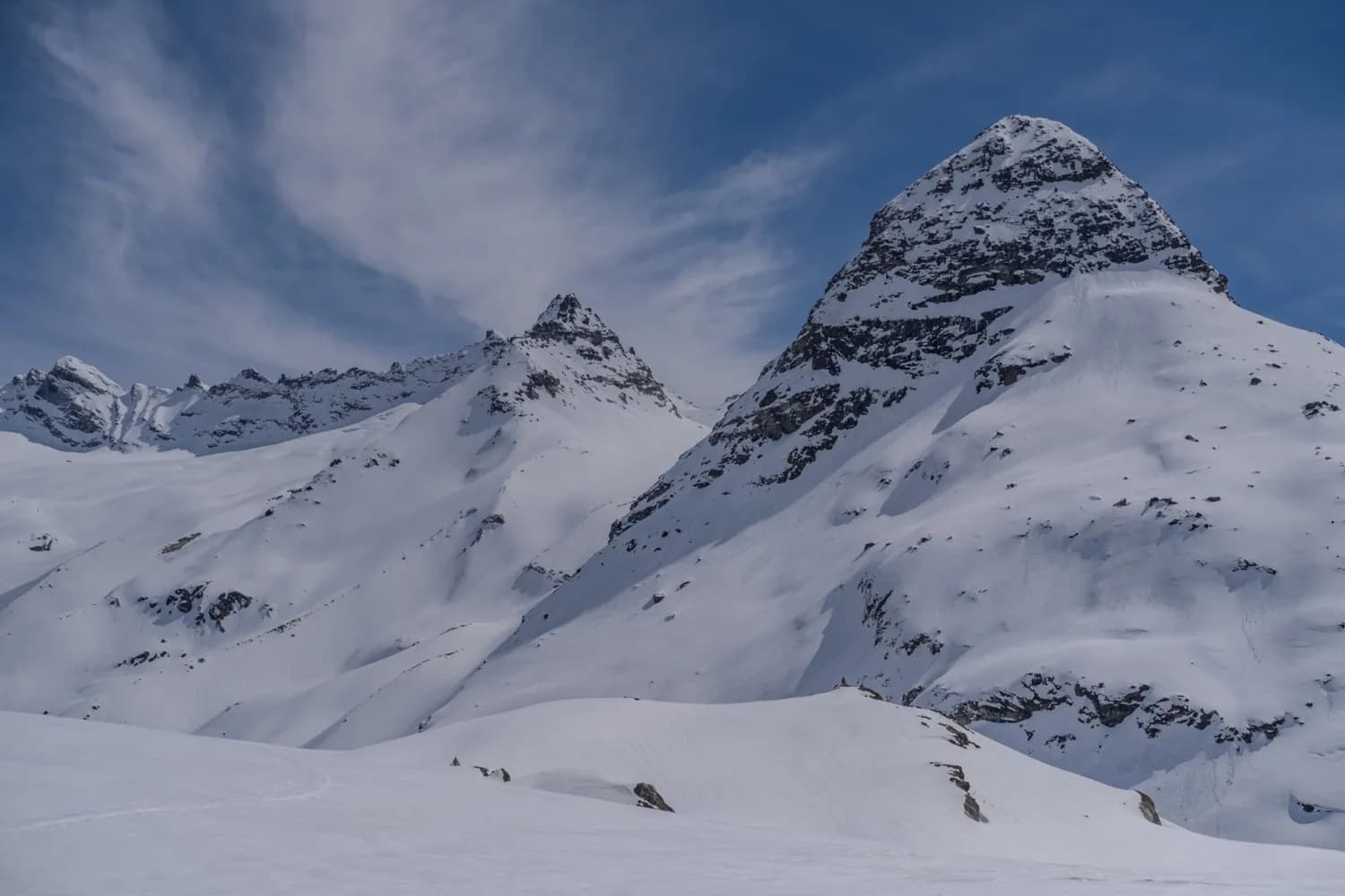 Snow-capped peaks rise majestically beneath a blue sky dotted with light clouds. The mountains, with their gentle yet steep slopes, are covered in a thick layer of snow.