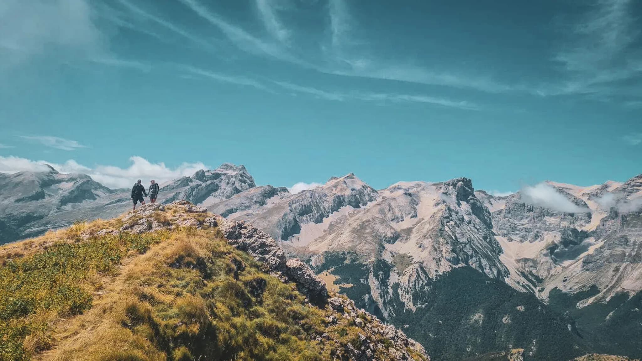 Two hikers hand in hand on a summit, with the majestic Alps in the background.