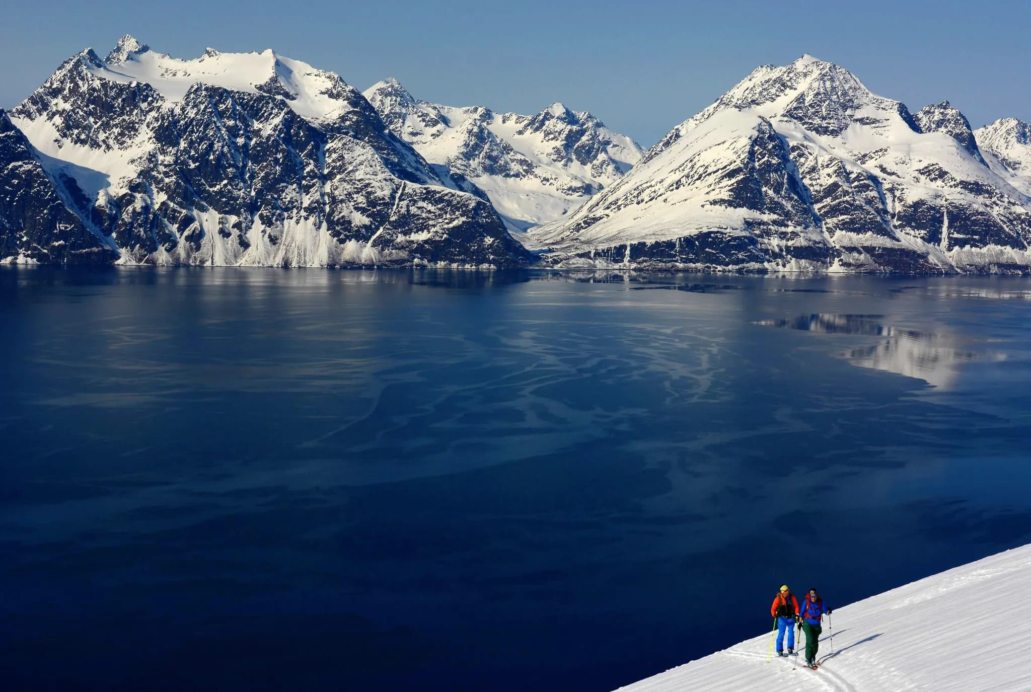 Un paysage montagneux spectaculaire avec des sommets enneigés surplombant un lac bleu profond. Deux personnes équipées pour une randonnée en montagne progressent lentement sur une pente recouverte de neige, tandis que la lumière du soleil éclaire