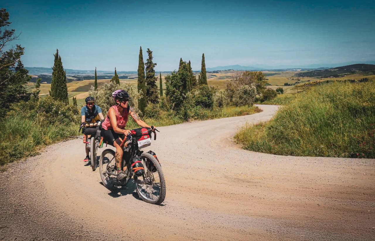 A pair of cyclists ride along a winding gravel road, surrounded by lush greenery and rolling hills. One of the cyclists, wearing a pink jersey and helmet.