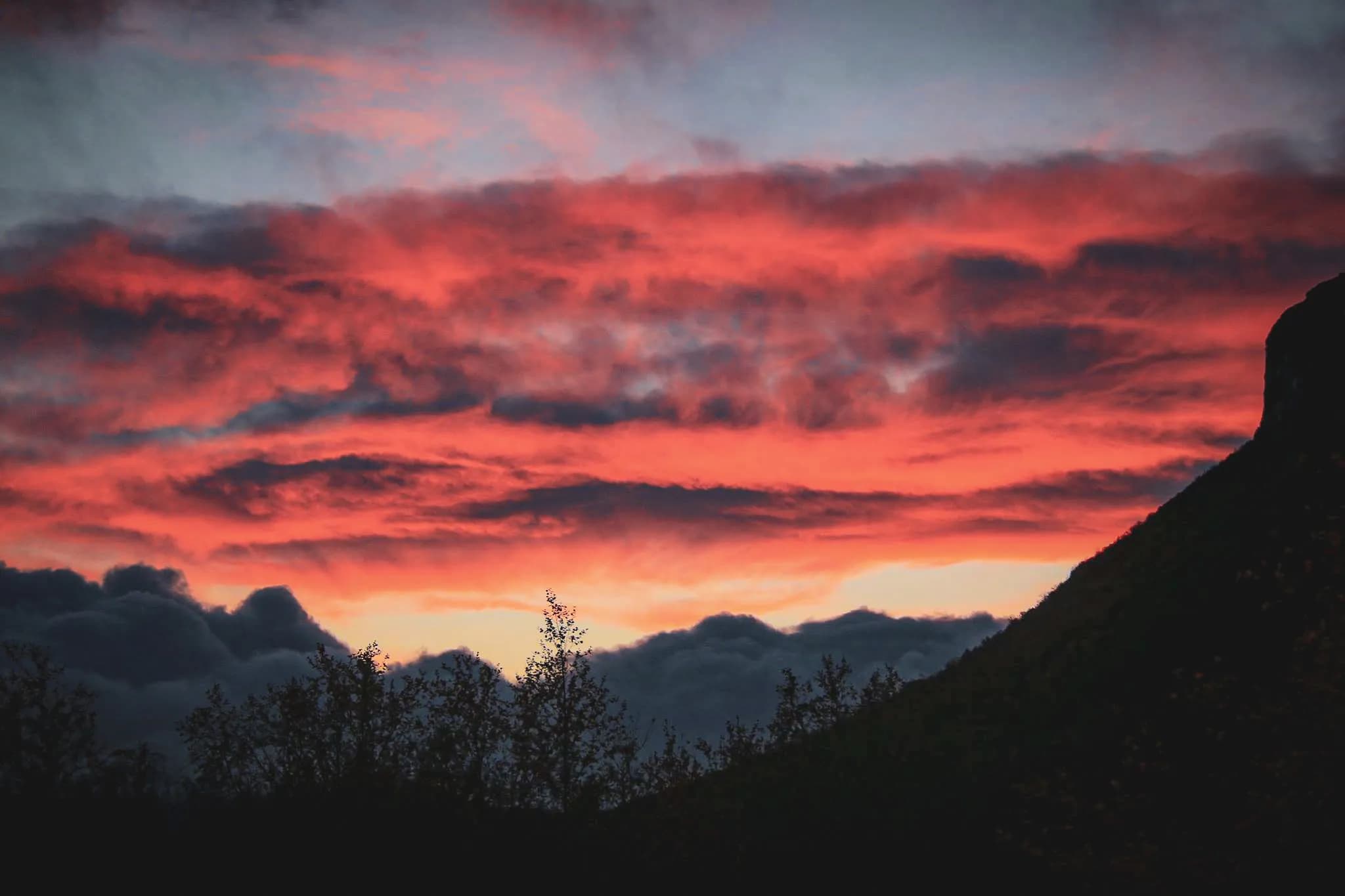 A vibrant sunset with shades of red, orange and blue, creating a striking contrast with the dark clouds. The silhouette of a mountain can be seen on the right, while trees and bushes can be seen on the left.