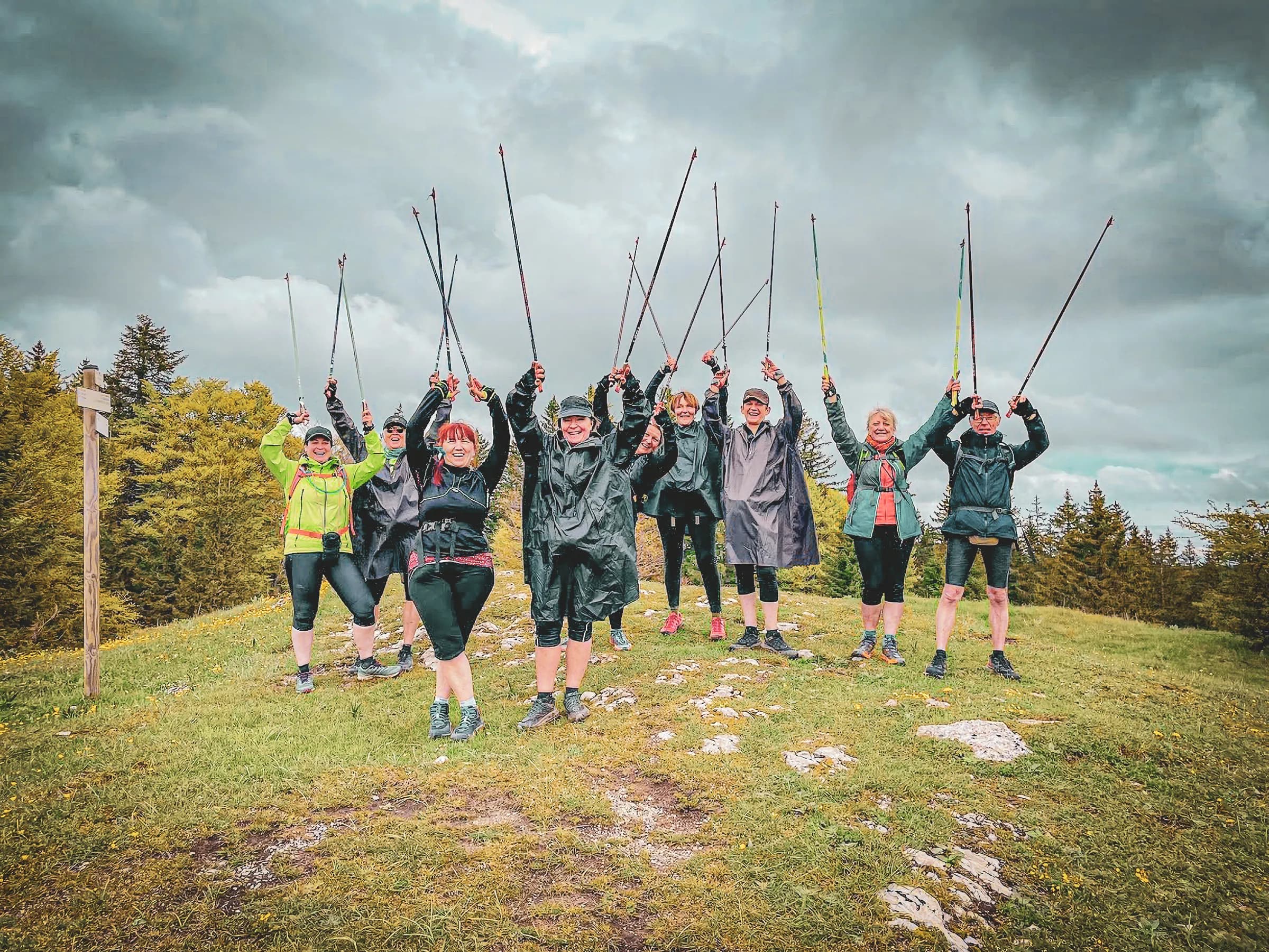 A group of eight people, dressed in hiking gear, stand on a green hill, raising their walking sticks in the air. Most of the participants are wearing waterproof clothing, and some are smiling happily.