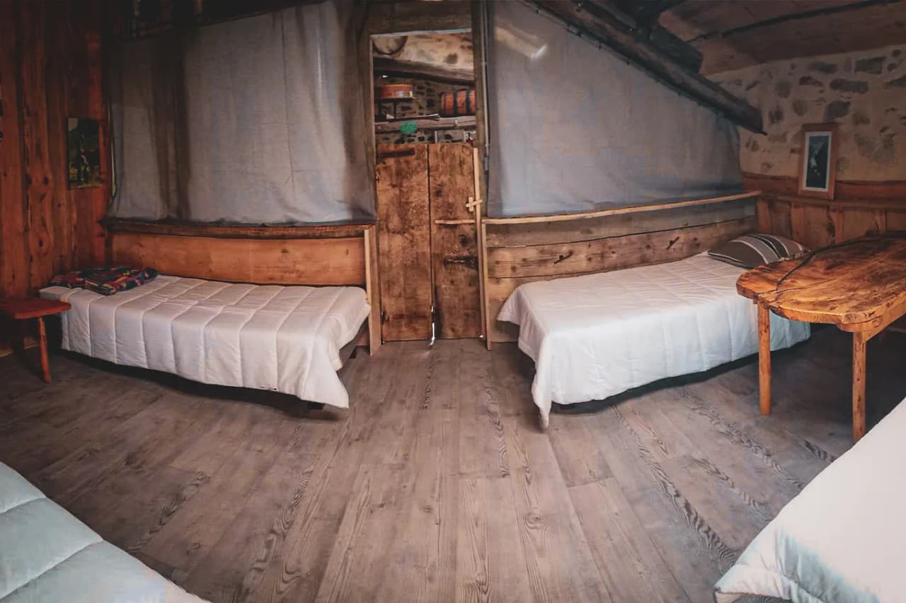A single room with three twin beds, each with a white cover. The walls are of wood and stone, creating a warm, rustic atmosphere. A solid wood door stands in the centre, leading to the bedroom.