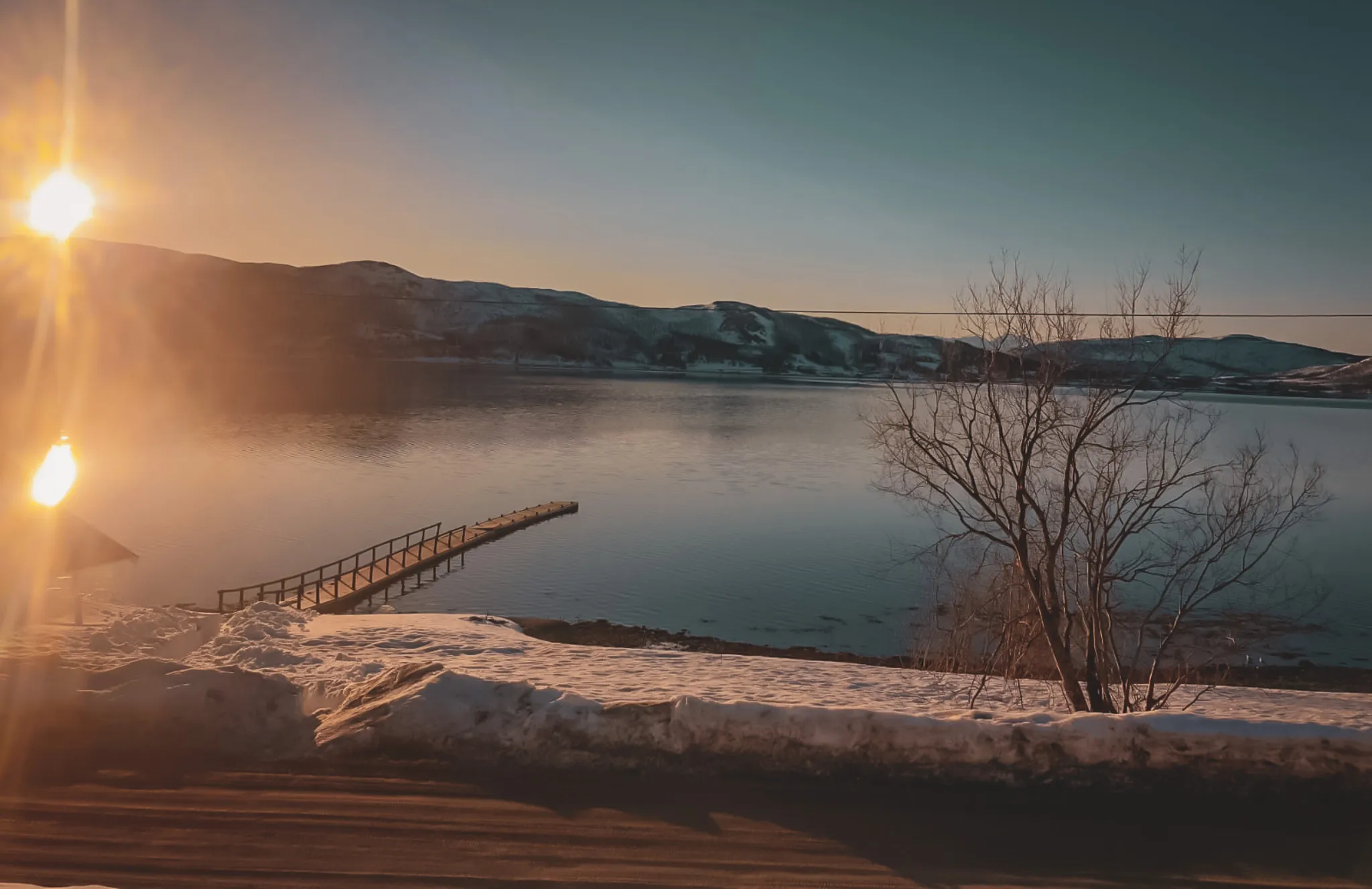Een rustig landschap bij zonsopgang, met een rustig meer dat de bergen in de verte weerspiegelt. Een kleine houten steiger steekt uit in het water, terwijl rechts een kale boom staat. De scène is omringd door