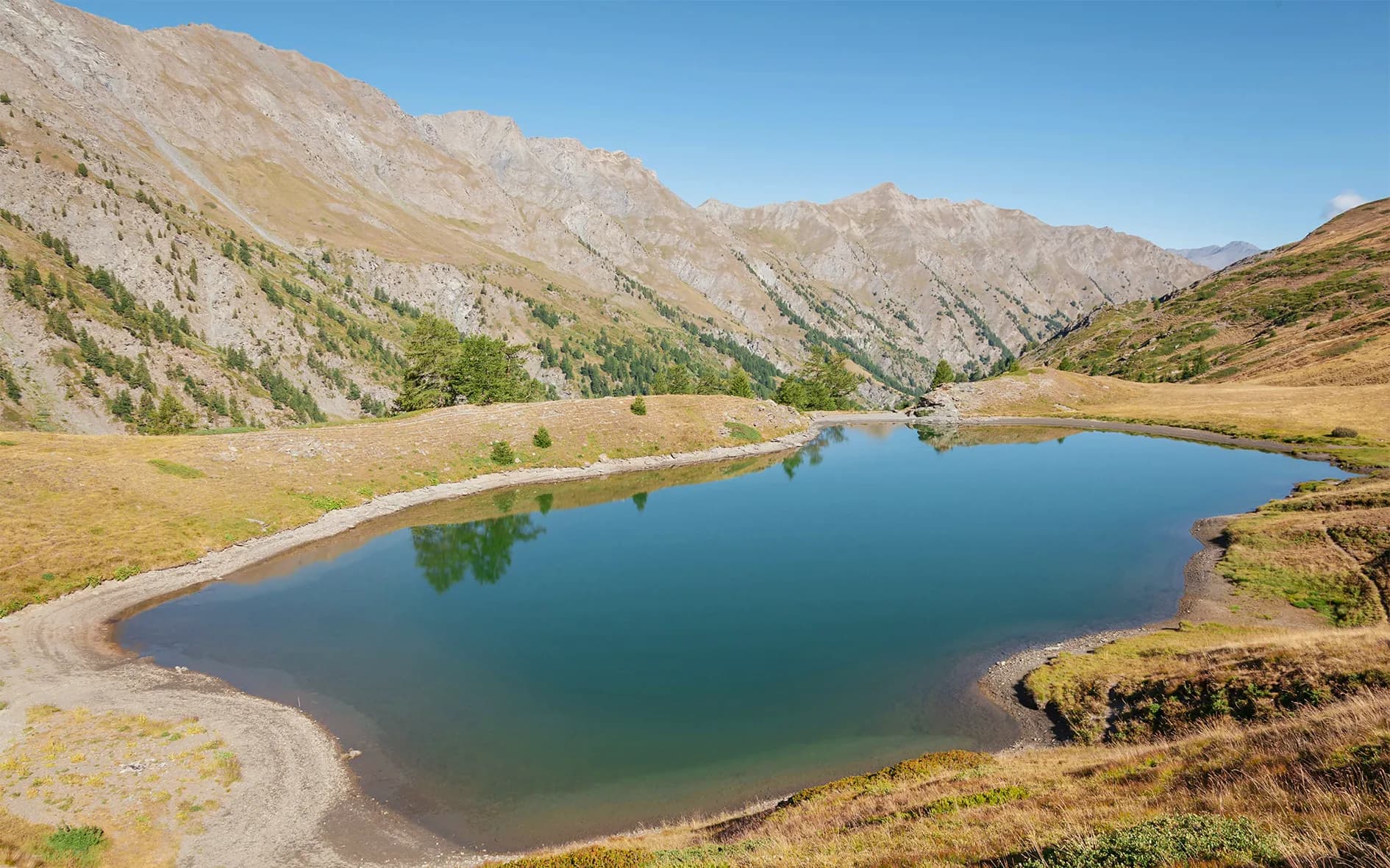 A peaceful lake surrounded by majestic mountains, with grassy slopes and pine forests on its sides. The surface of the water is calm, reflecting the clear blue sky and surrounding landscapes.