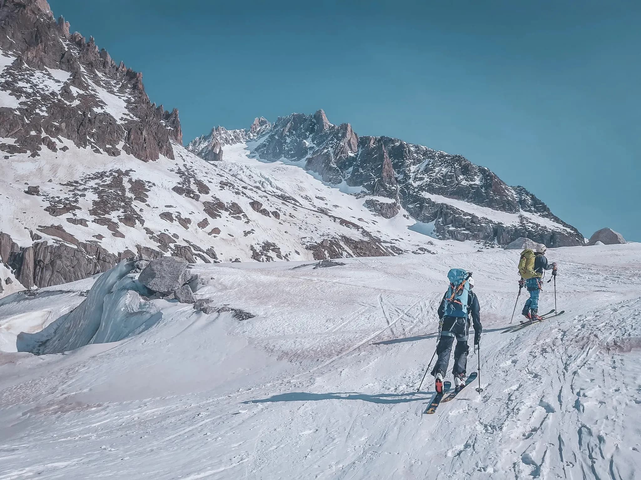 Deux skieurs progressent sur un glacier, entourés de montagnes majestueuses sous un ciel bleu.