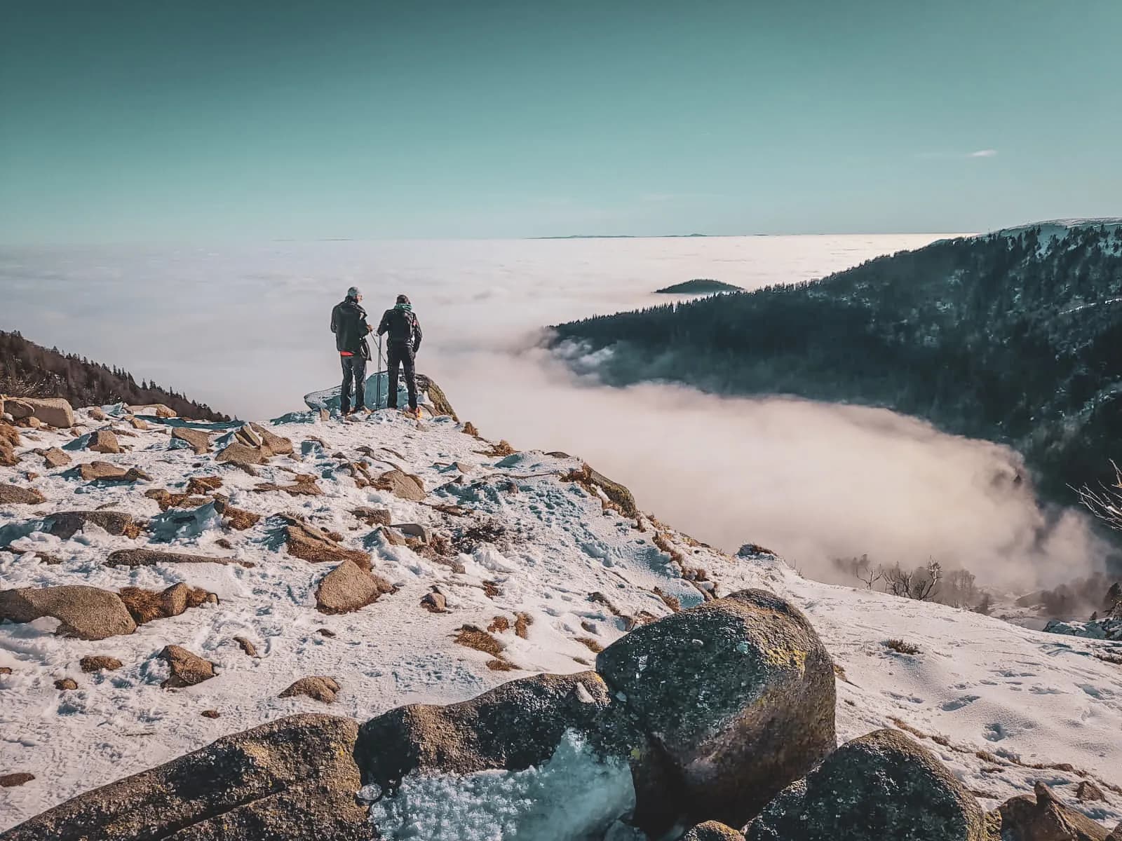 Een besneeuwd berglandschap met uitzicht op een zee van wolken. Twee mensen, uitgerust om te wandelen, staan aan de rand van een rotsachtige bergkam en bewonderen het panoramische uitzicht. Op