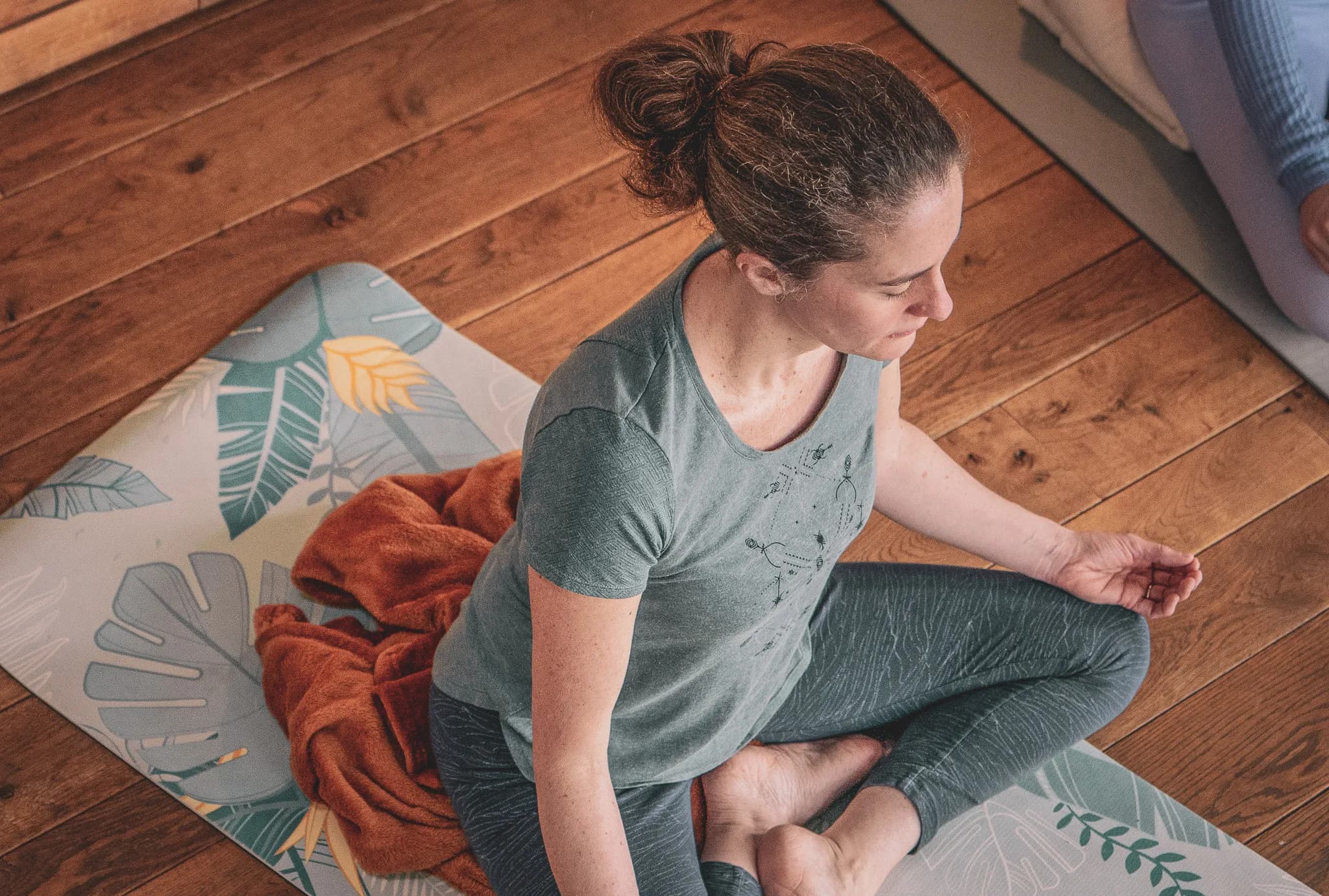 A woman is practising meditation in a calm environment. She is sitting on a yoga mat on a wooden floor, wearing a grey t-shirt and matching leggings. An orange plaid is wrapped around her. L