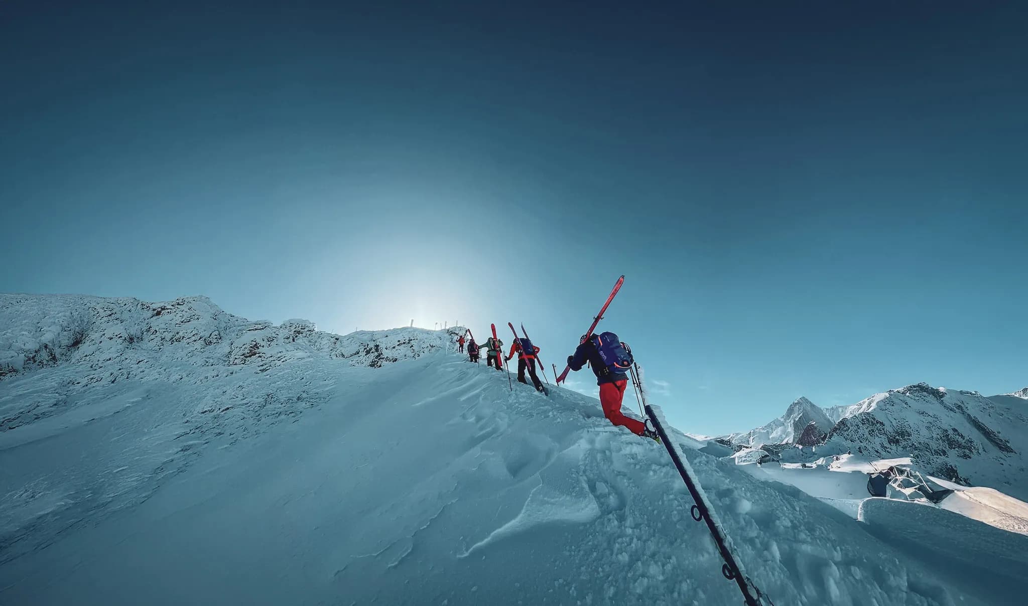 A group of skiers in the mountains, making their way up a snowy slope. The sky is clear, with soft light coming from the background, suggesting a time close to sunrise or sunset.