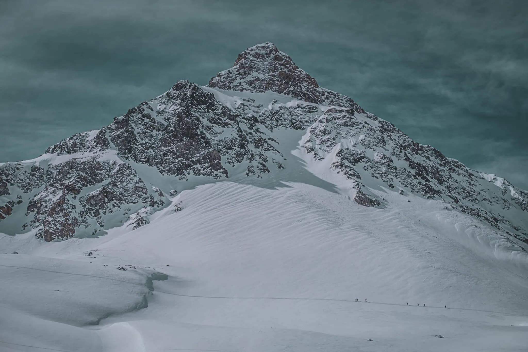A majestic snow-capped mountain peak, with sheer cliffs and a jagged ridge. In the foreground, icy slopes stretch out, dotted with natural landforms and tracks left by the snow.