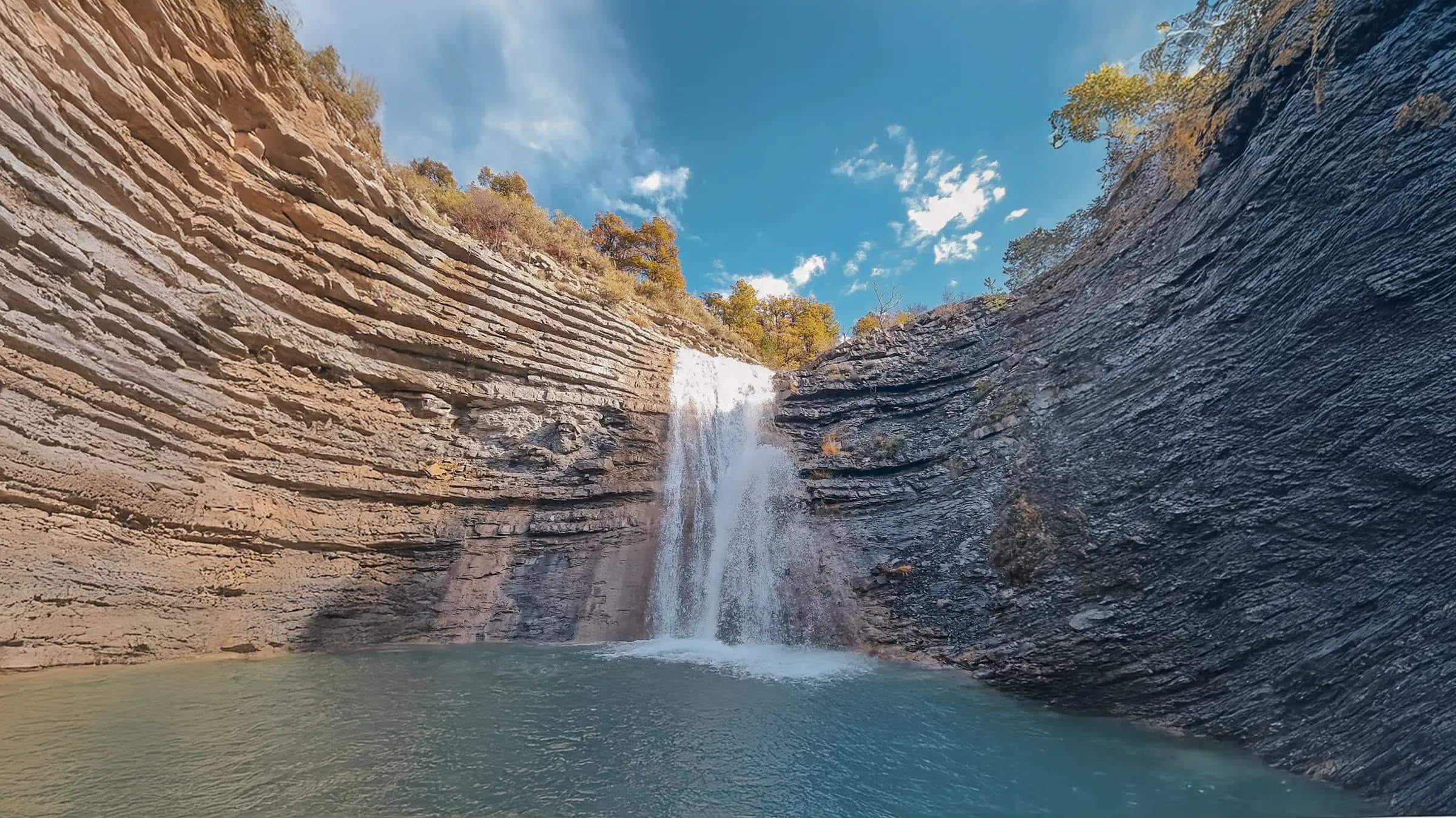 A majestic waterfall cascades over layered rocks, creating a pool of clear water below. The cliffs surrounding the waterfall have distinct layers of stone that add character to the scene. The vegetation