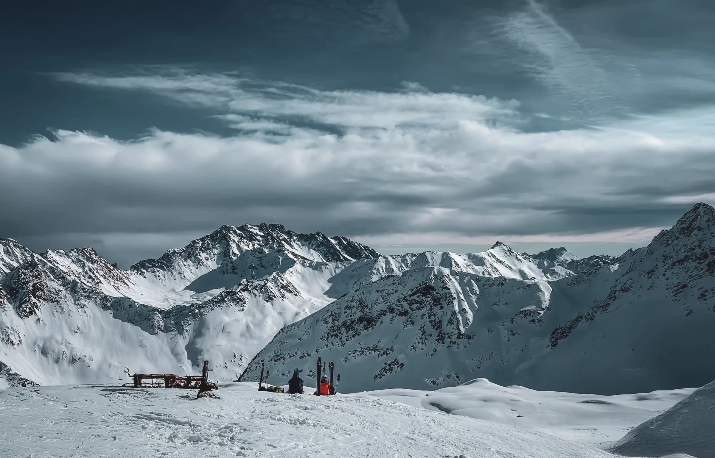 A snowy mountain landscape, with majestic peaks covered in snow. In the foreground, a few people are sitting on the snow, accompanied by skis stuck in the ground. The sky is cloudy, with