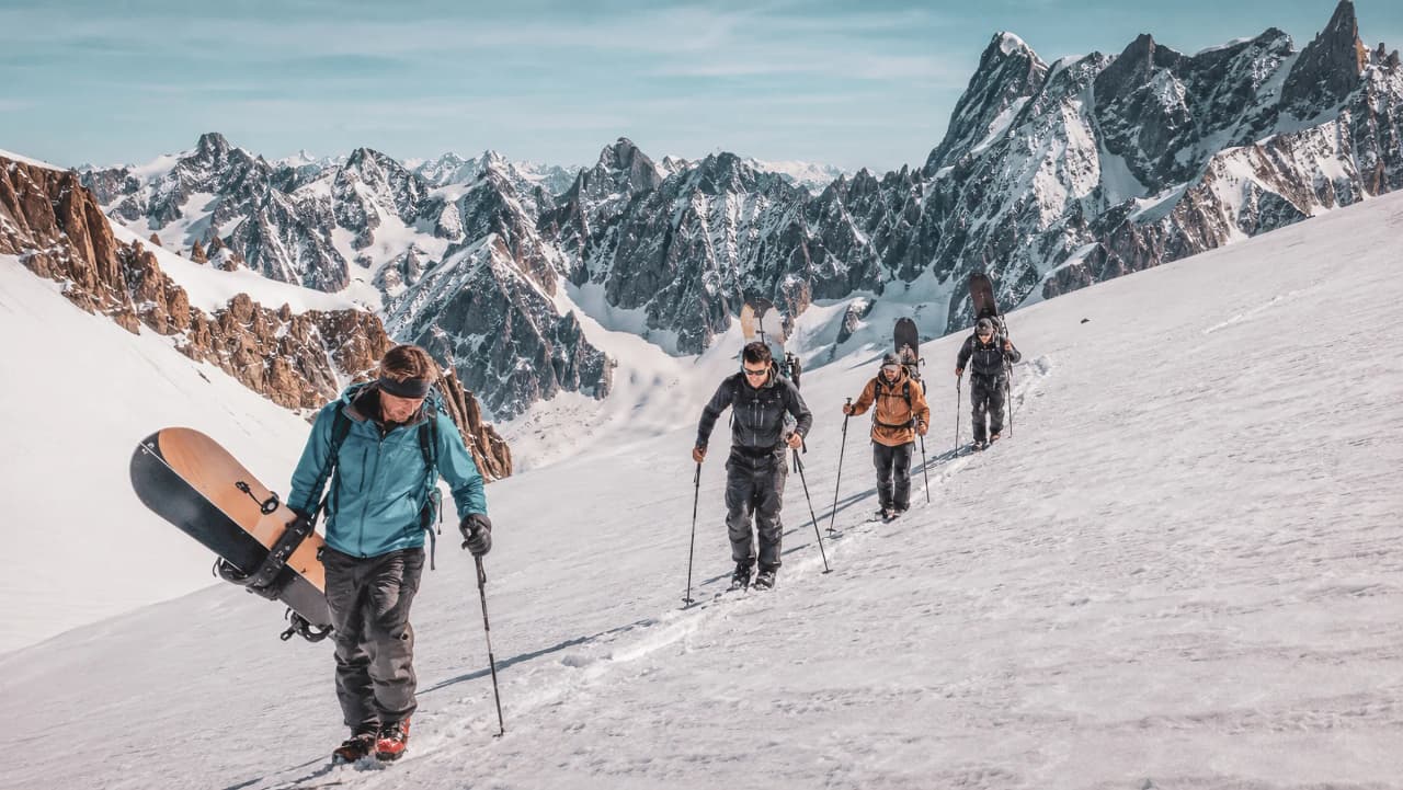 Groupe de snowboardeurs en pleine ascension sur un glacier, avec des pics majestueux en arrière-plan.