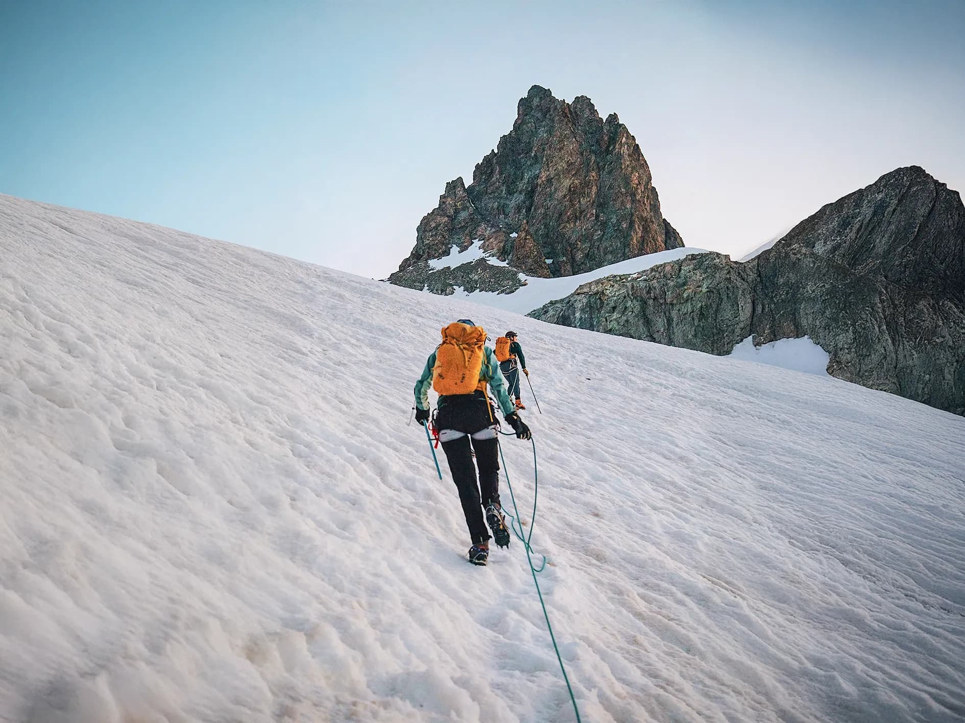 A group of hikers climbing a snowy slope. The mountaineers are wearing bright orange rucksacks and clinging to a rope as they make their way towards a majestic rocky peak that looms behind them.