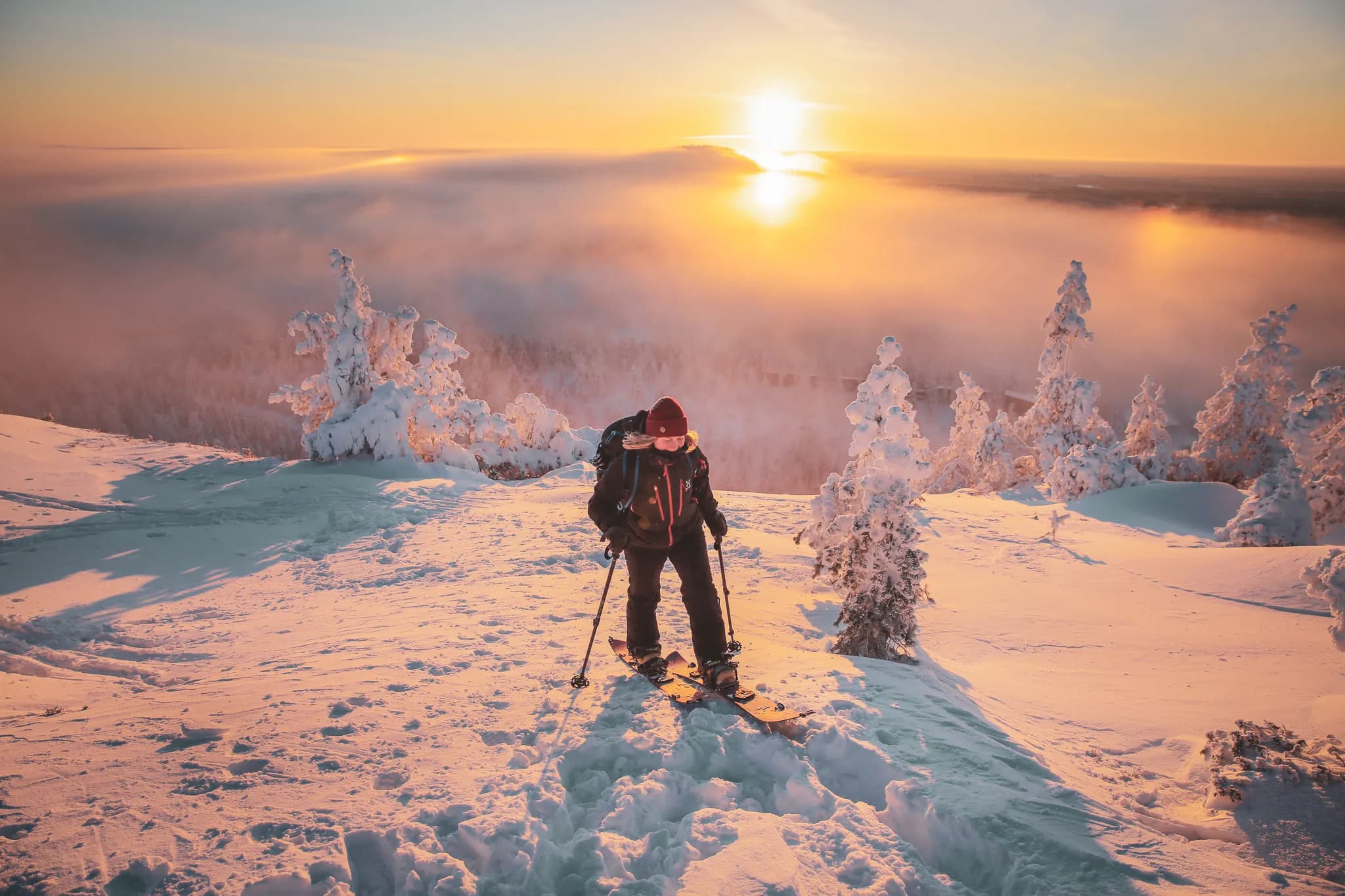 A skier stands on a snow-covered slope, surrounded by snow-covered fir trees. The sunrise casts golden and pink hues across the sky, while a light mist envelops the valley.