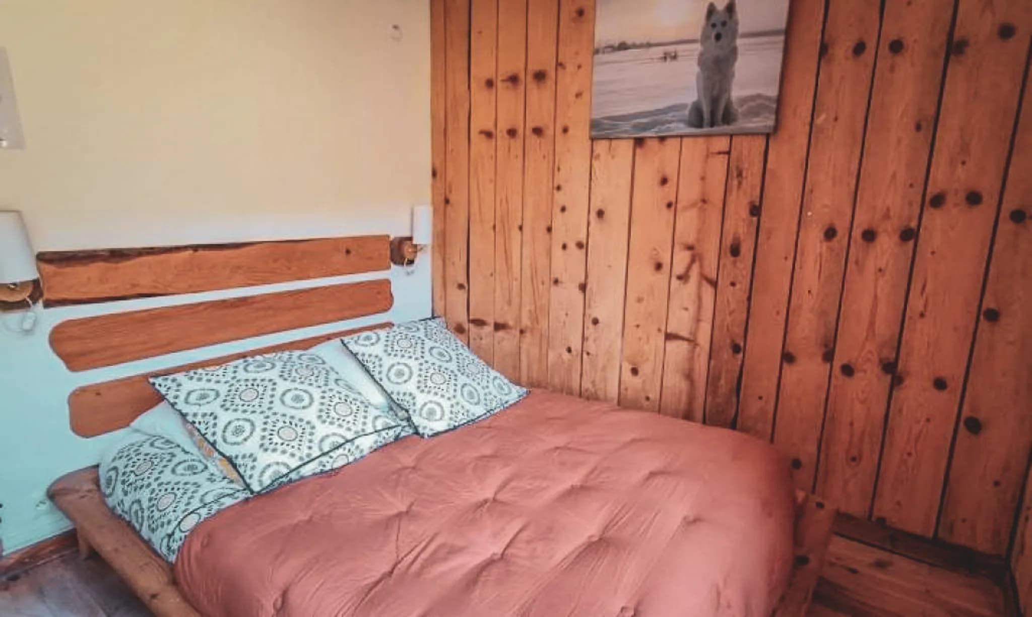 A cosy bedroom with a wooden bed and a headboard made from natural wood planks. The cushions have a geometric pattern, and the bed is covered with a pink quilt.