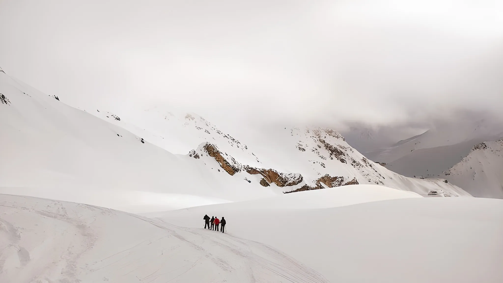 Vijf sneeuwschoenwandelaars doorkruisen een besneeuwd landschap in de richting van majestueuze bergtoppen.