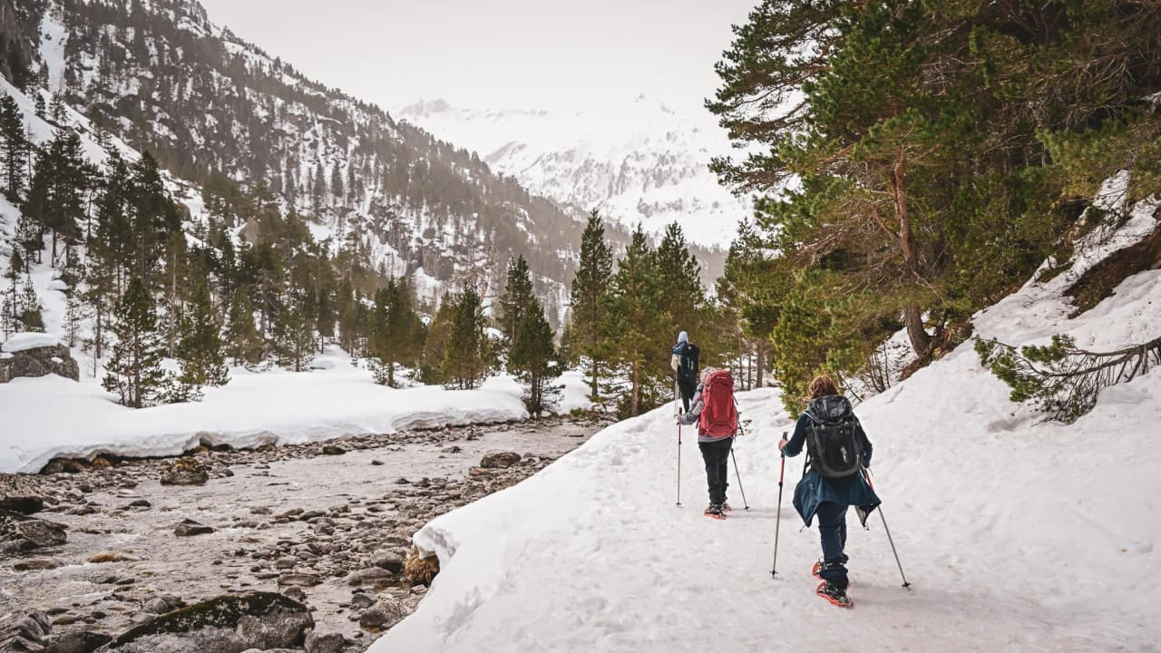 Three hikers on snowshoes walk along a river in a snowy landscape in the Pyrenees.