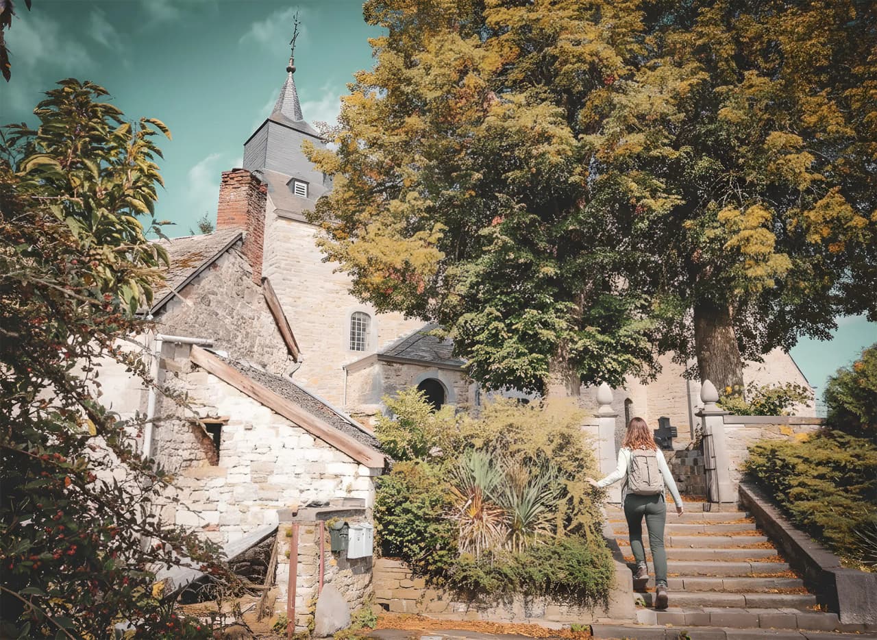 Une randonneuse gravit des escaliers bordés de verdure, découvrant un charmant village en Ardenne.