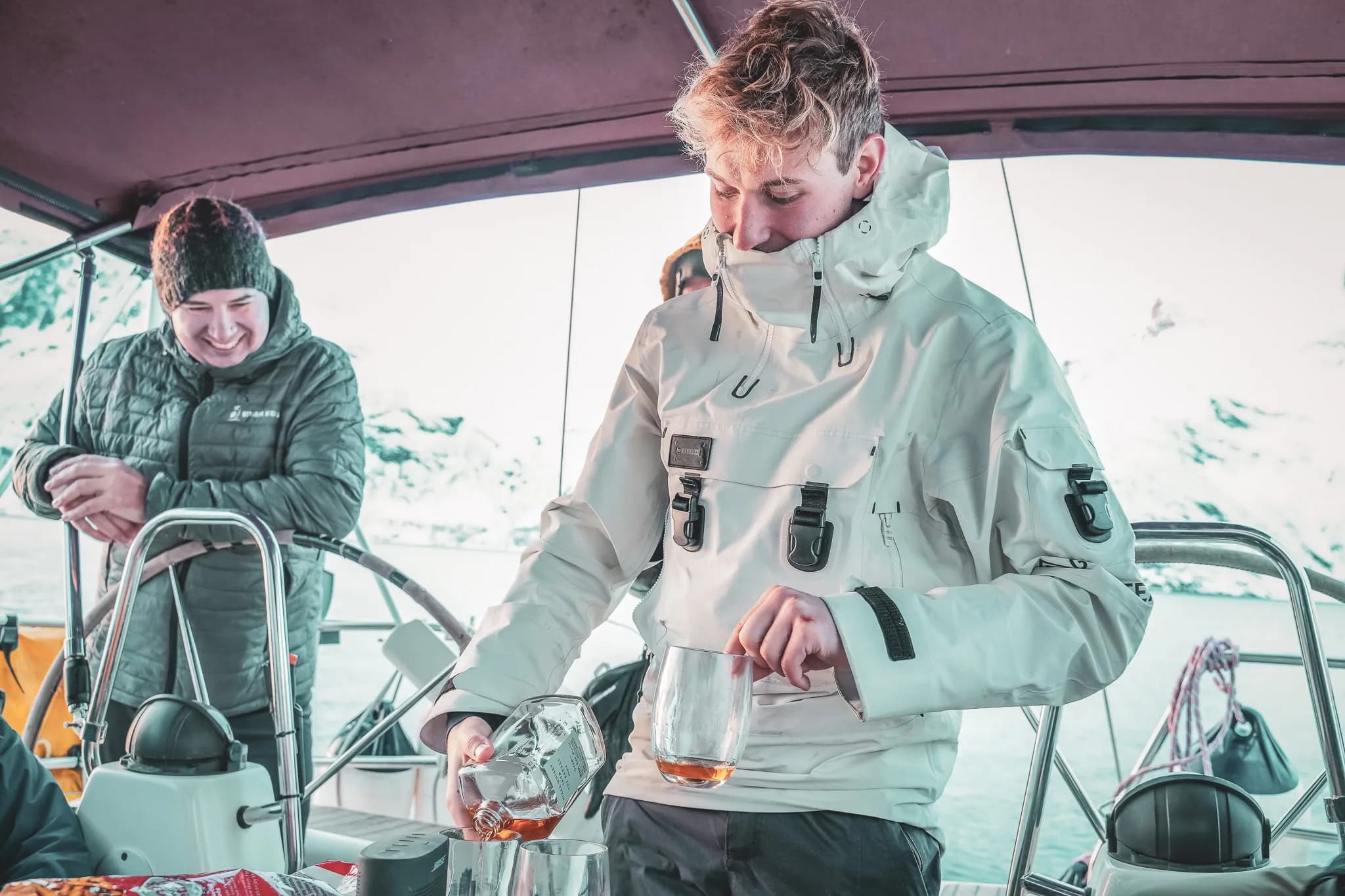 Un jeune homme en manteau blanc verse une boisson dans un verre sur un bateau, tandis qu'un autre homme, vêtu d'une veste noire et d'un bonnet, sourit en arrière-plan. L'ambiance semble détendue, et le