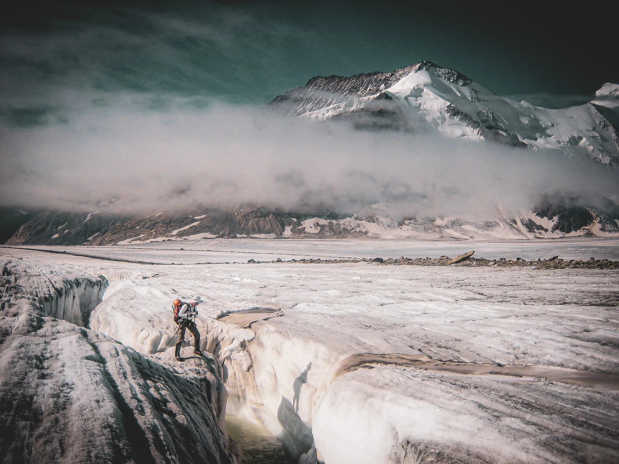 A snow-covered mountain icefield with a hiker standing on a point of ice, overlooking a crevasse. In the background, steep peaks.