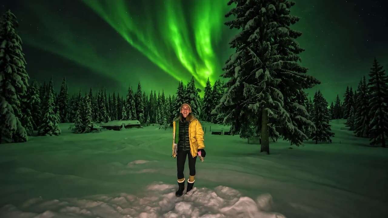 Femme souriante sous une magnifique aurore boréale, entourée de sapins enneigés en Laponie.
