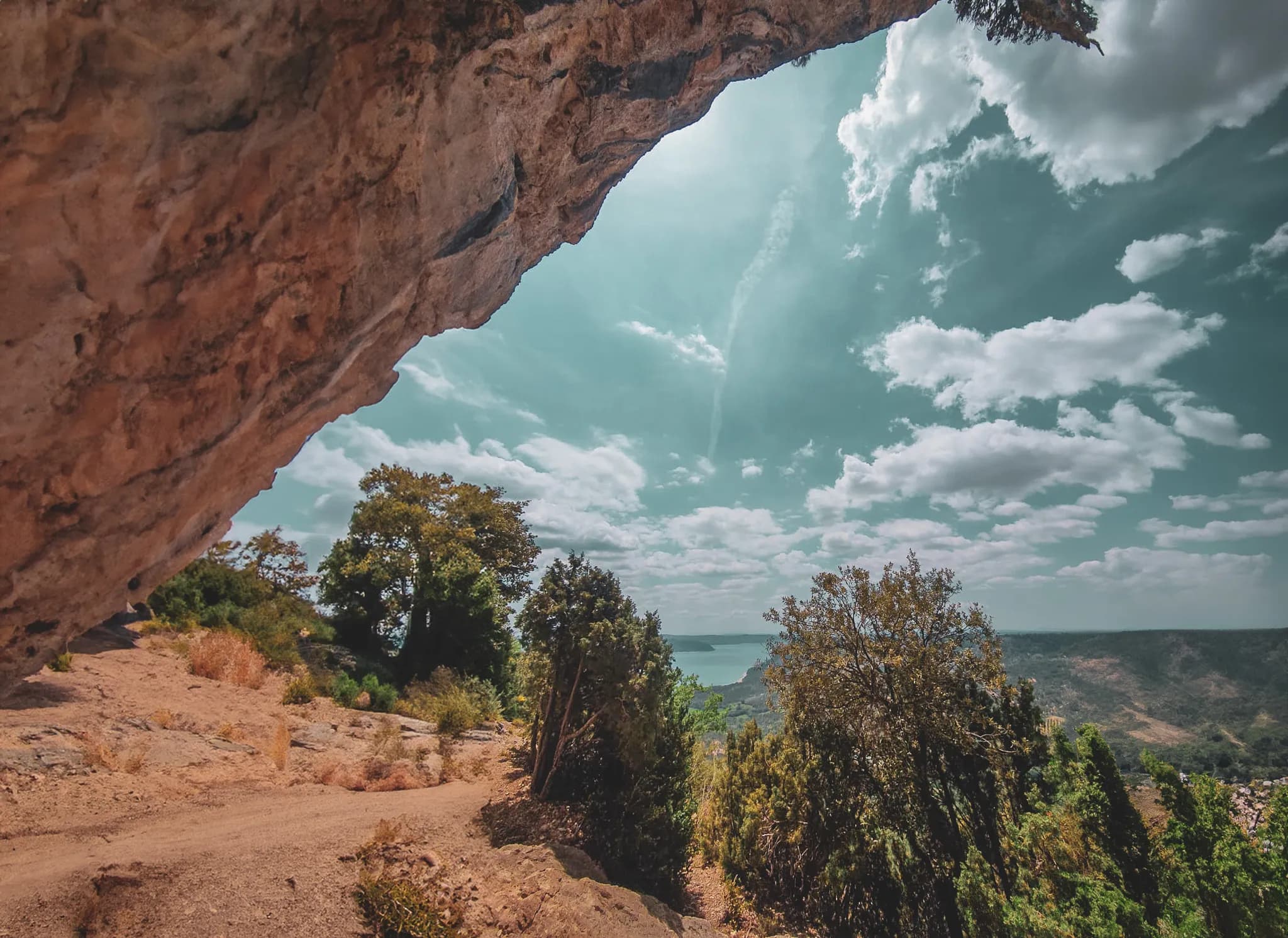 A spectacular natural landscape, with an unobstructed view of a green ravine. In the foreground, a massive rock formation dominates the space, while a winding path crosses the scene, lined with bushes and trees.